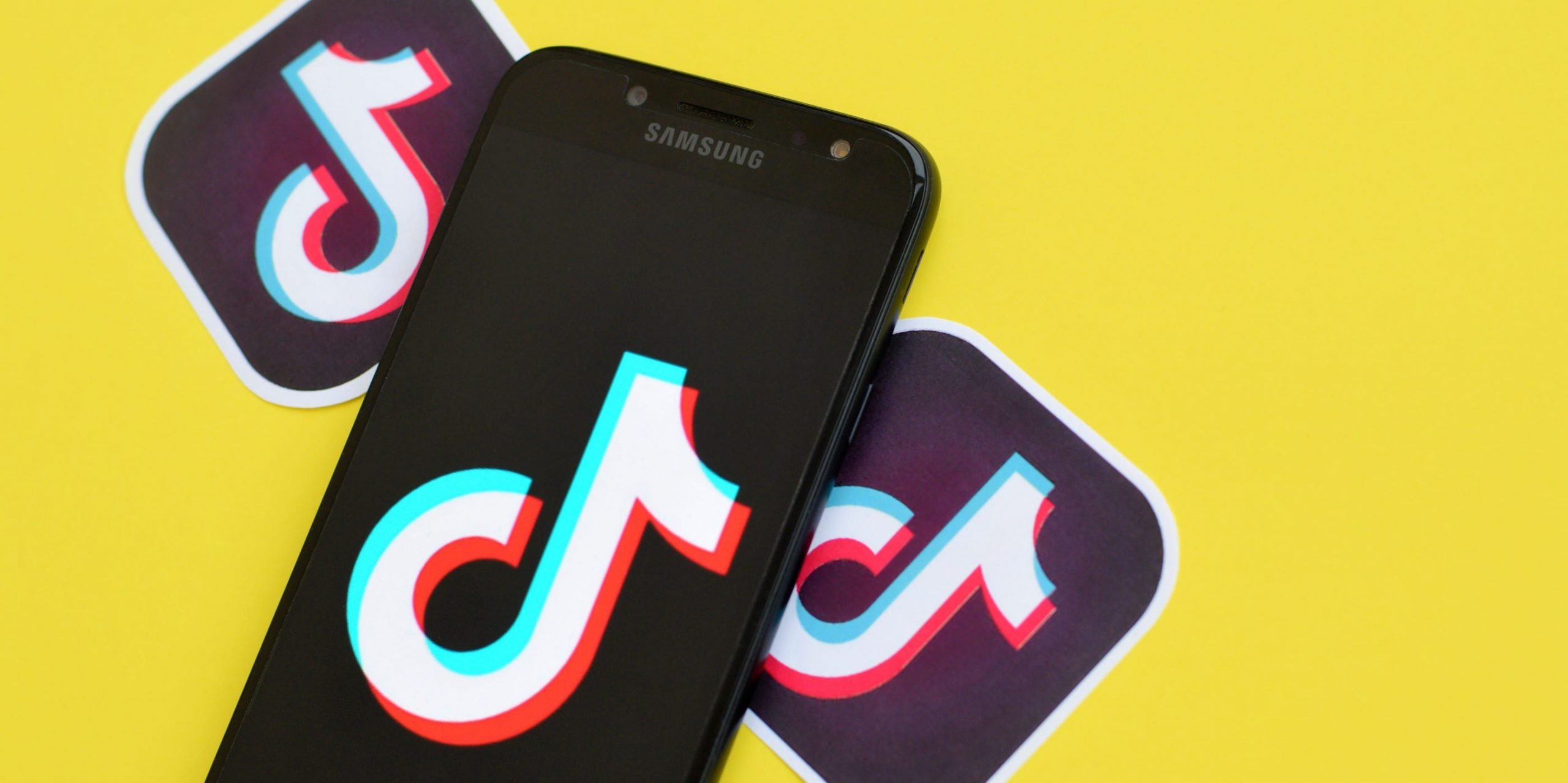 A Samsung phone displaying the TikTok logo, sitting in front of a yellow background.