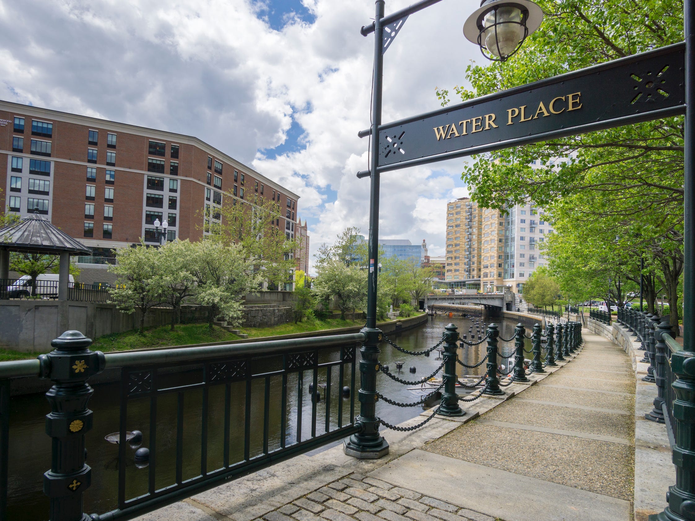 a sidewalk with a guardrail and a sign that says water place next to the a river with buildings in the background