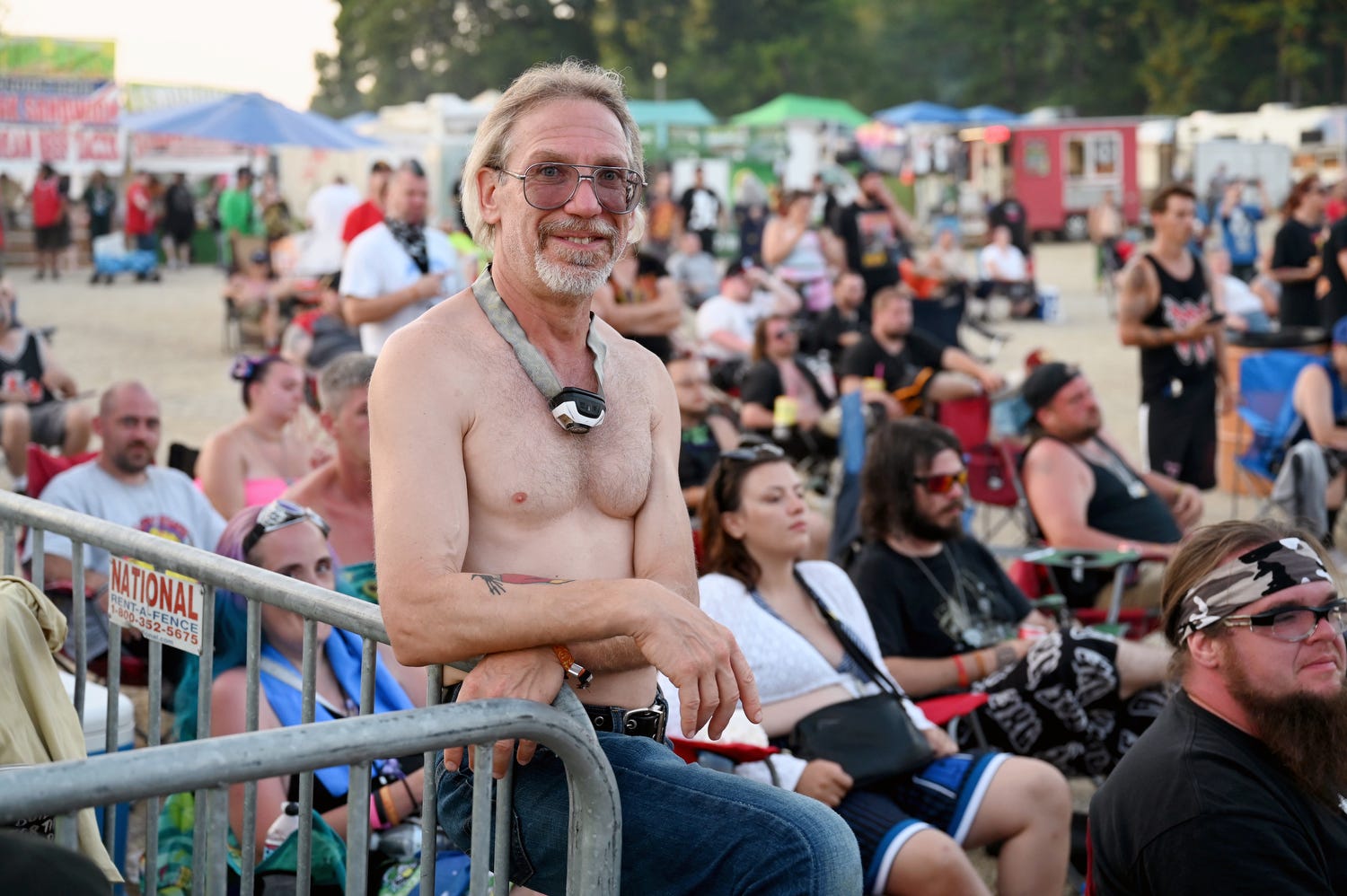 A shirtless male fan wearing glasses sits on a barricade with concertgoers sitting behind him.