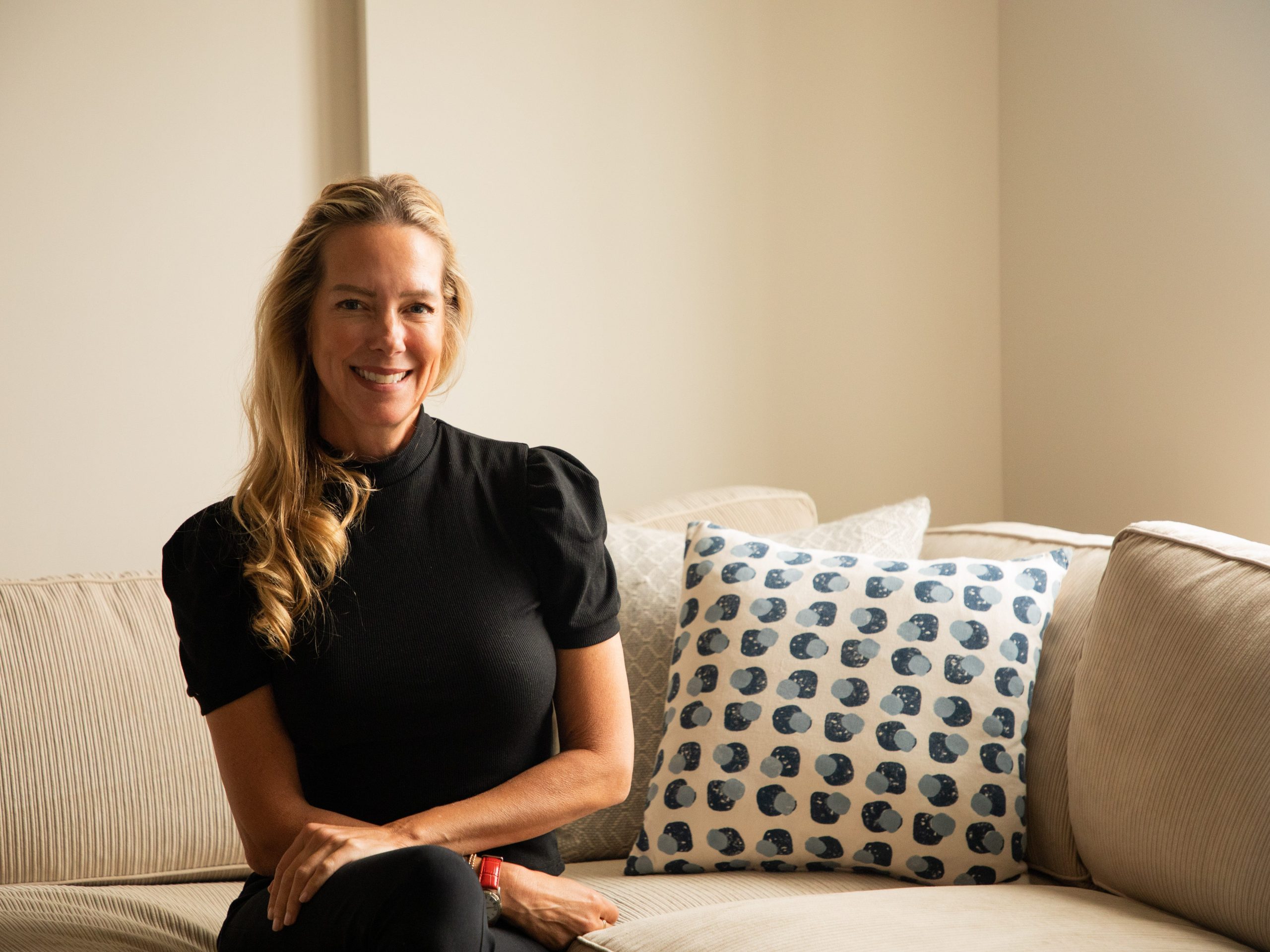 A woman with blonde hair sits on a couch and smiles.