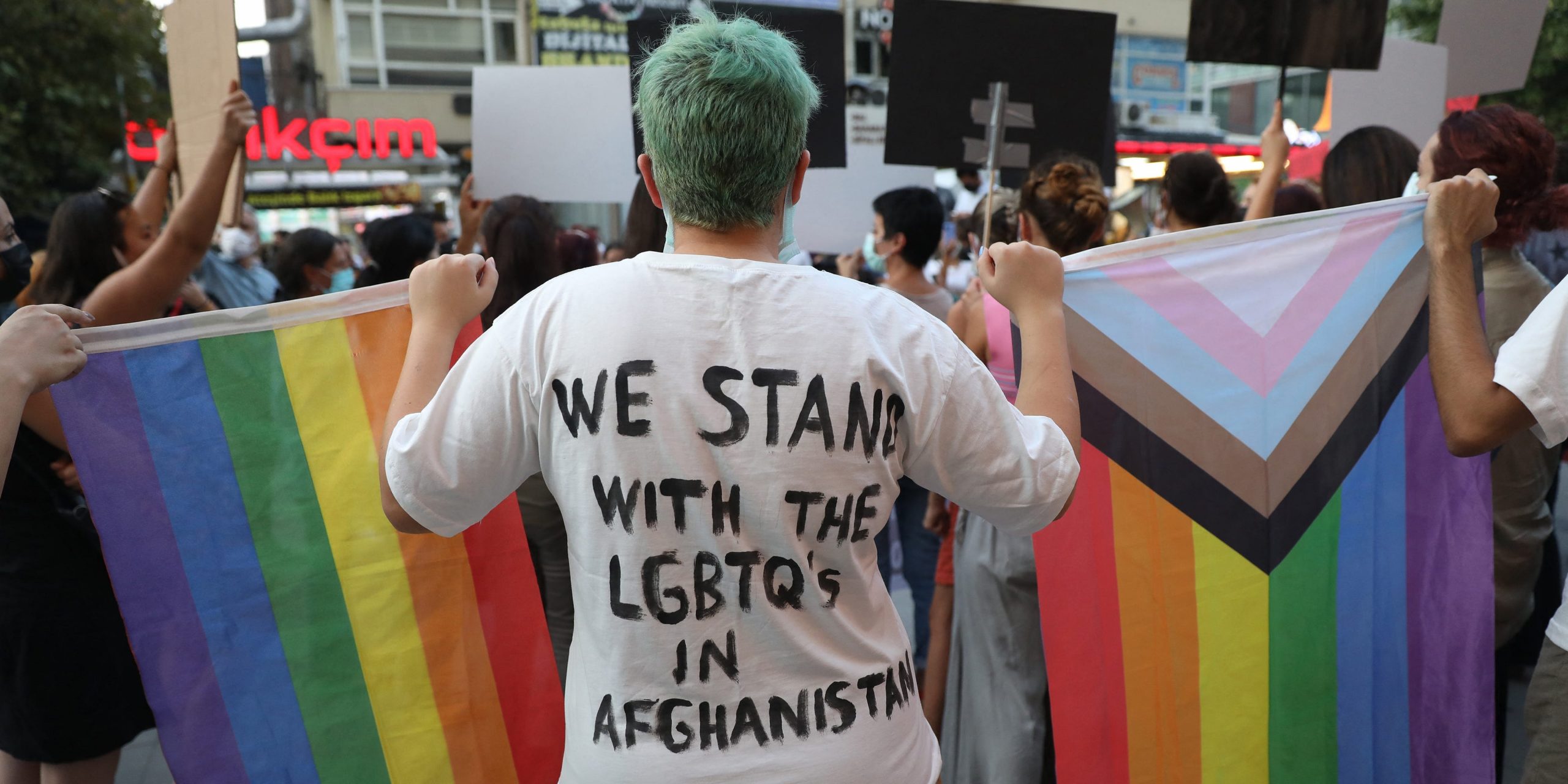 A person holds a rainbow flag and wears a shirt that says, "We stand with the LGBTQs in Afghanistan."