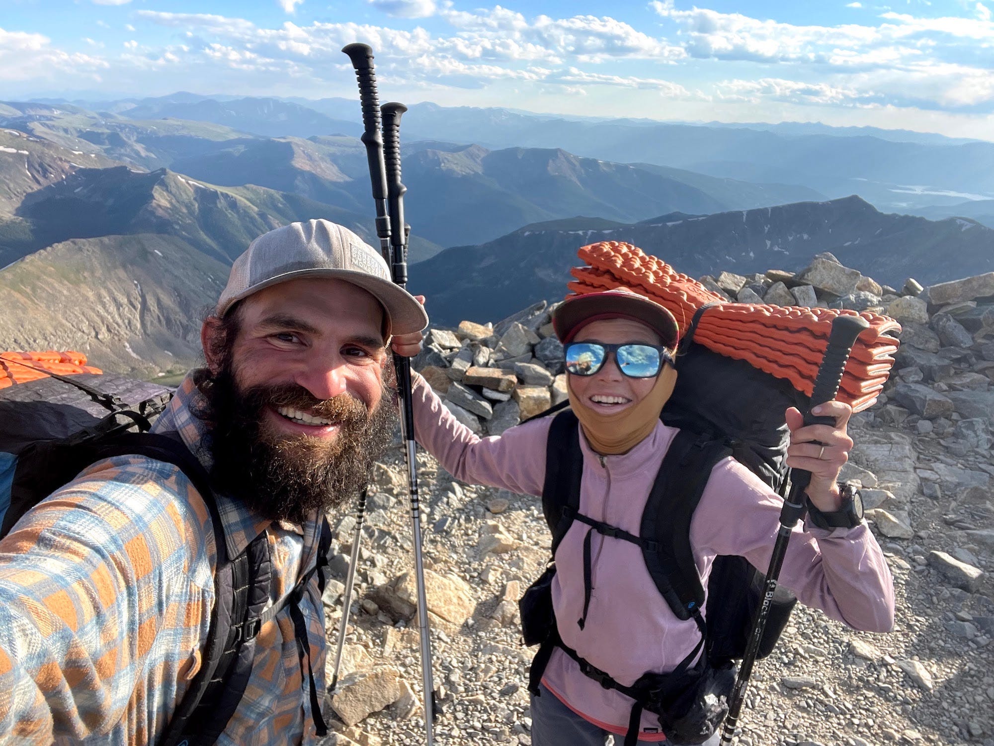 A couple smiles in hiking gear atop a mountain.