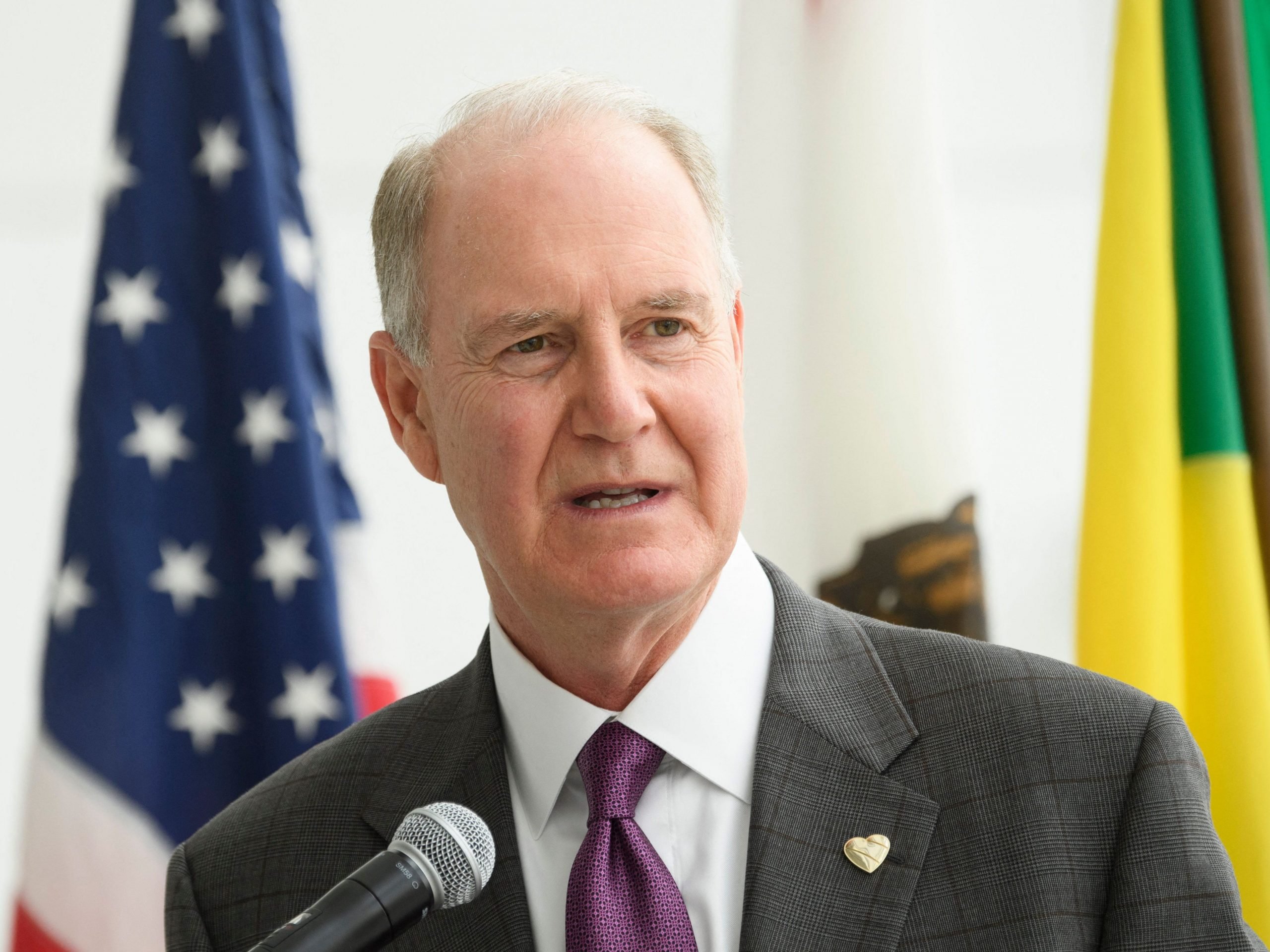 Gary Kelly wears a grey suit and purple tie while speaking into a microphone in front of a US flag.