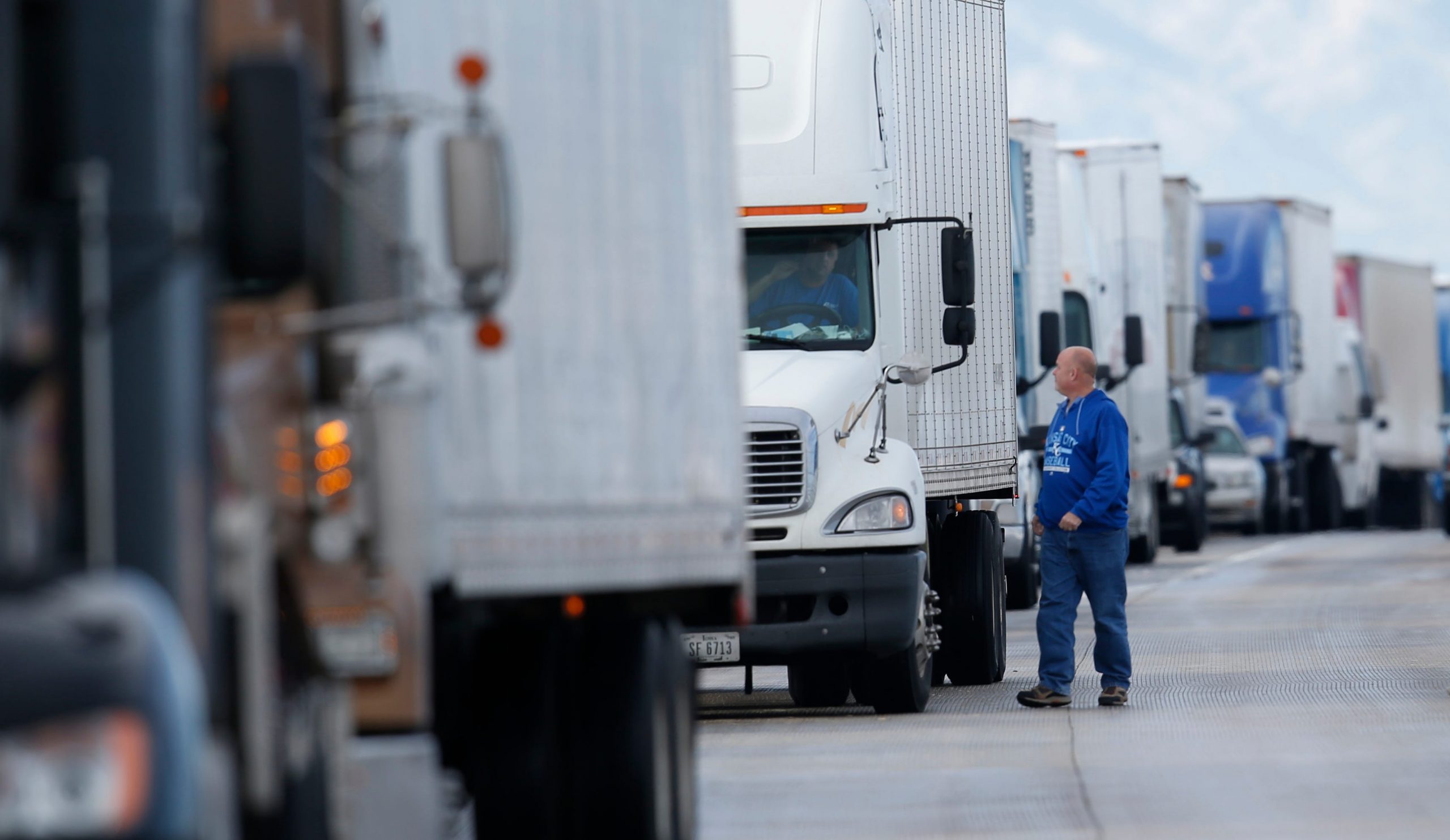 A man wearing a blue sweater walks next to a large white truck which part of a longer line of trucks.