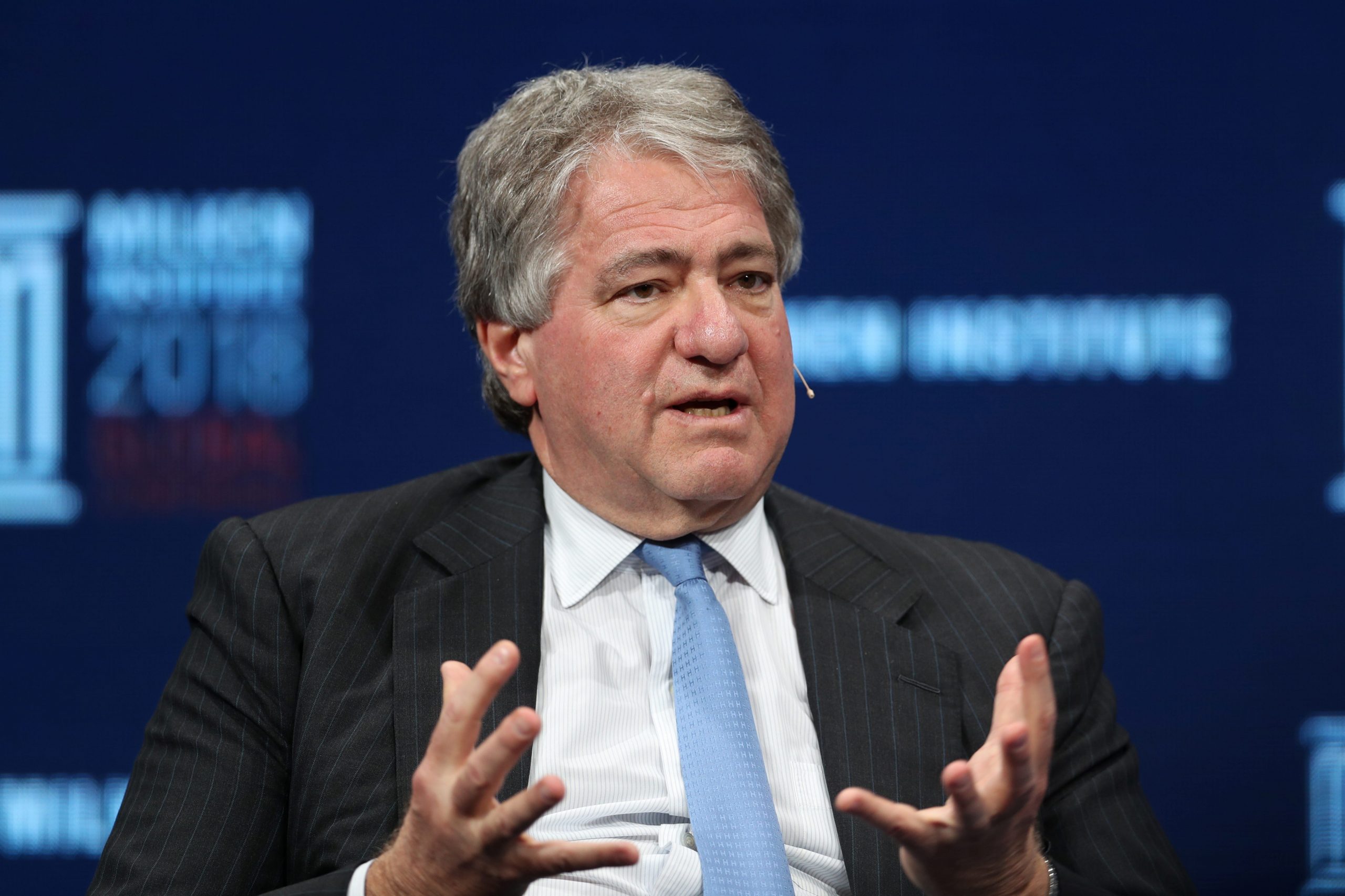 Leon Black wears a dark grey suit, white shirt, and blue tie while speaking on stage.