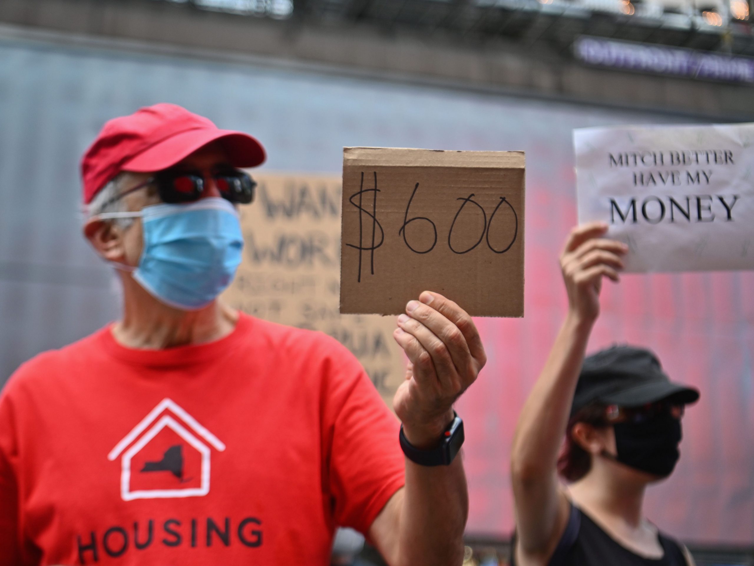 A man in a red shirt and hat and a blue face mask holds up a cardboard sign that says "$600."
