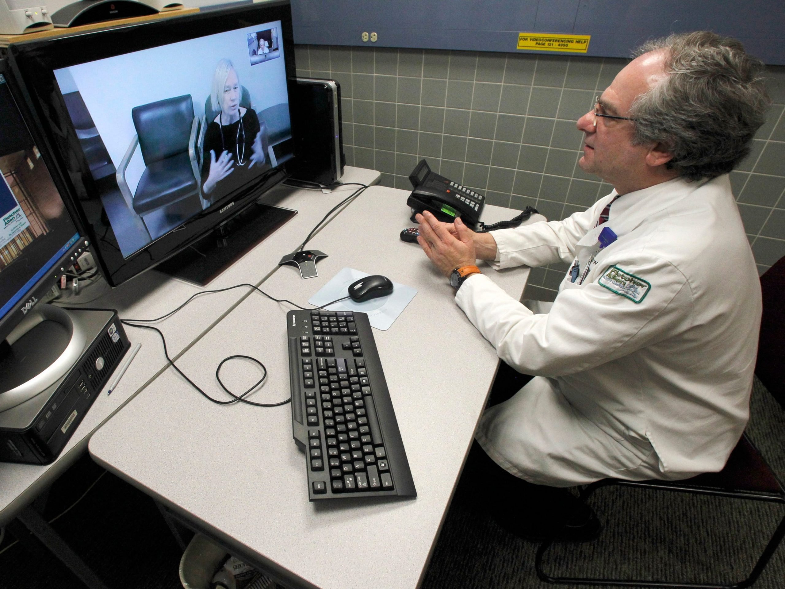 A doctor wearing a white lab coat sits at a desk in front of a monitor with a woman speaking on the screen.