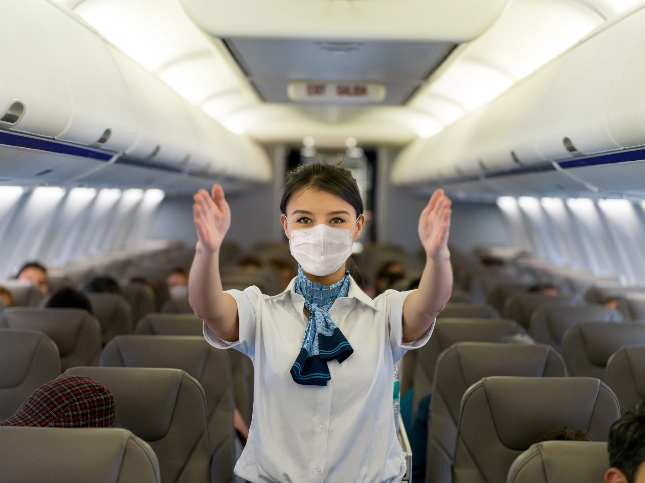 A woman flight attendants wearing a mask gestures towards the exits on an airplane.