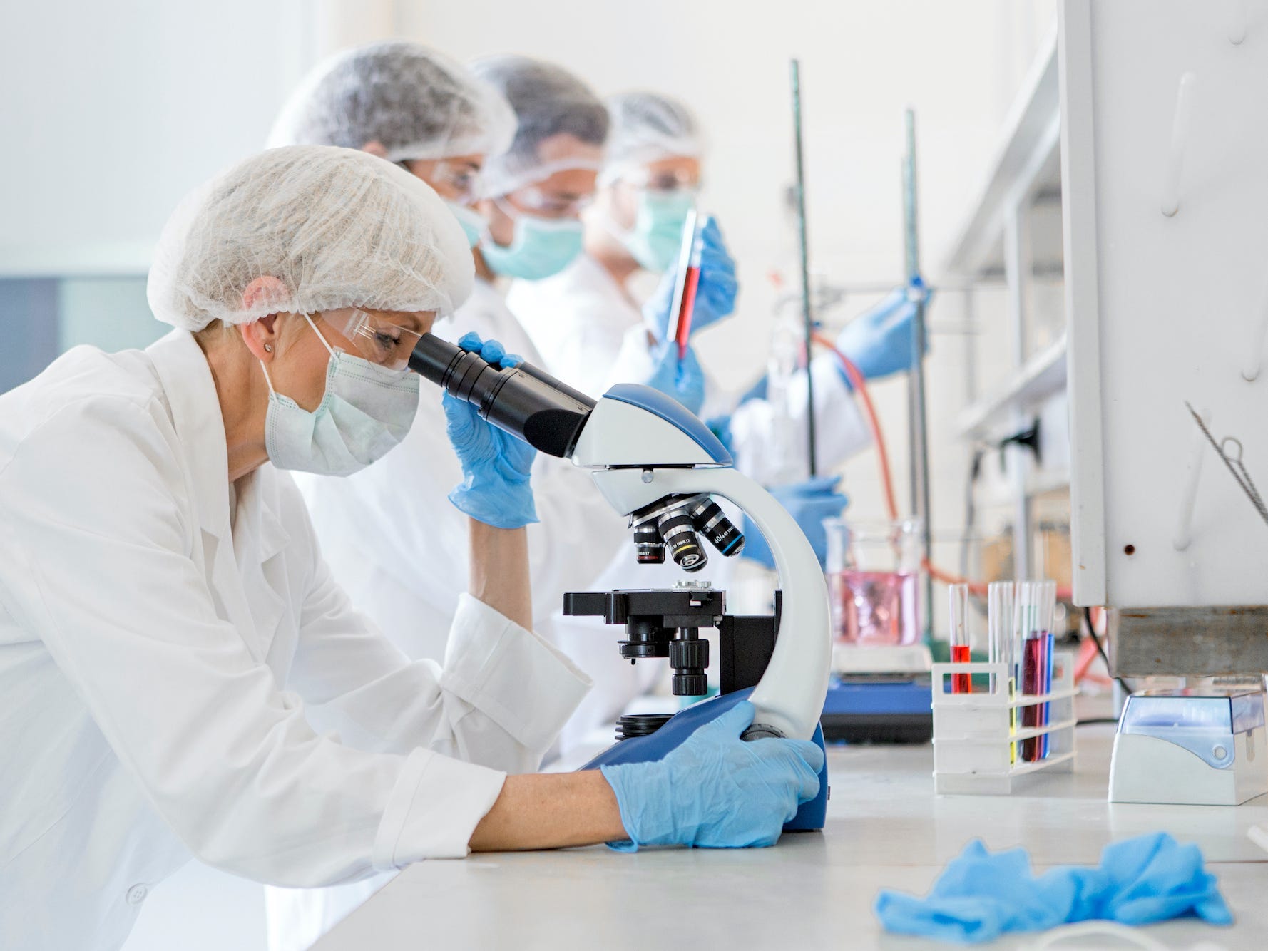 A scientist, who is wearing blue gloves and a lab coat, looks through a microscope. There are other scientists working next to her.