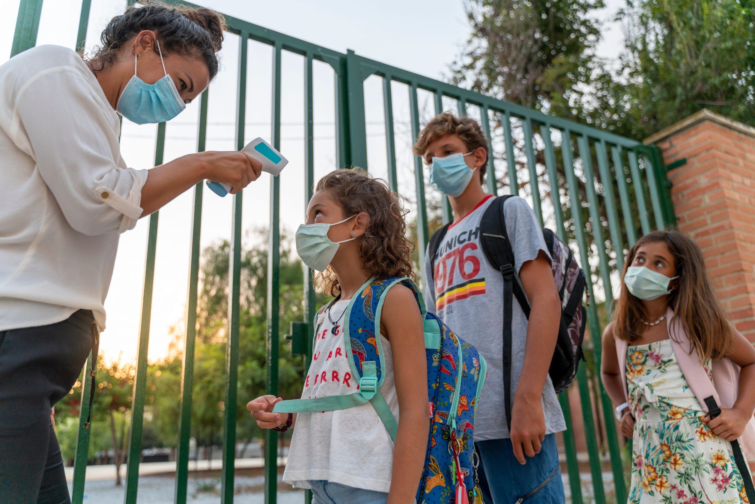 A teacher takes the temperature of pupils lining up outside school wearing face masks.