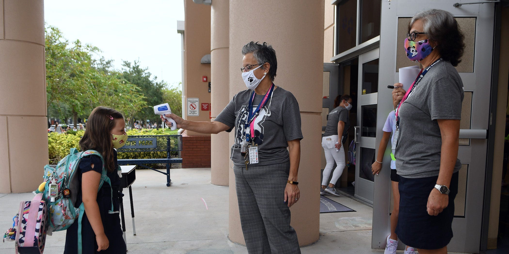 A schoolteacher, wearing a mask, checks the temperature of one of her masked students, before the child is let inside.