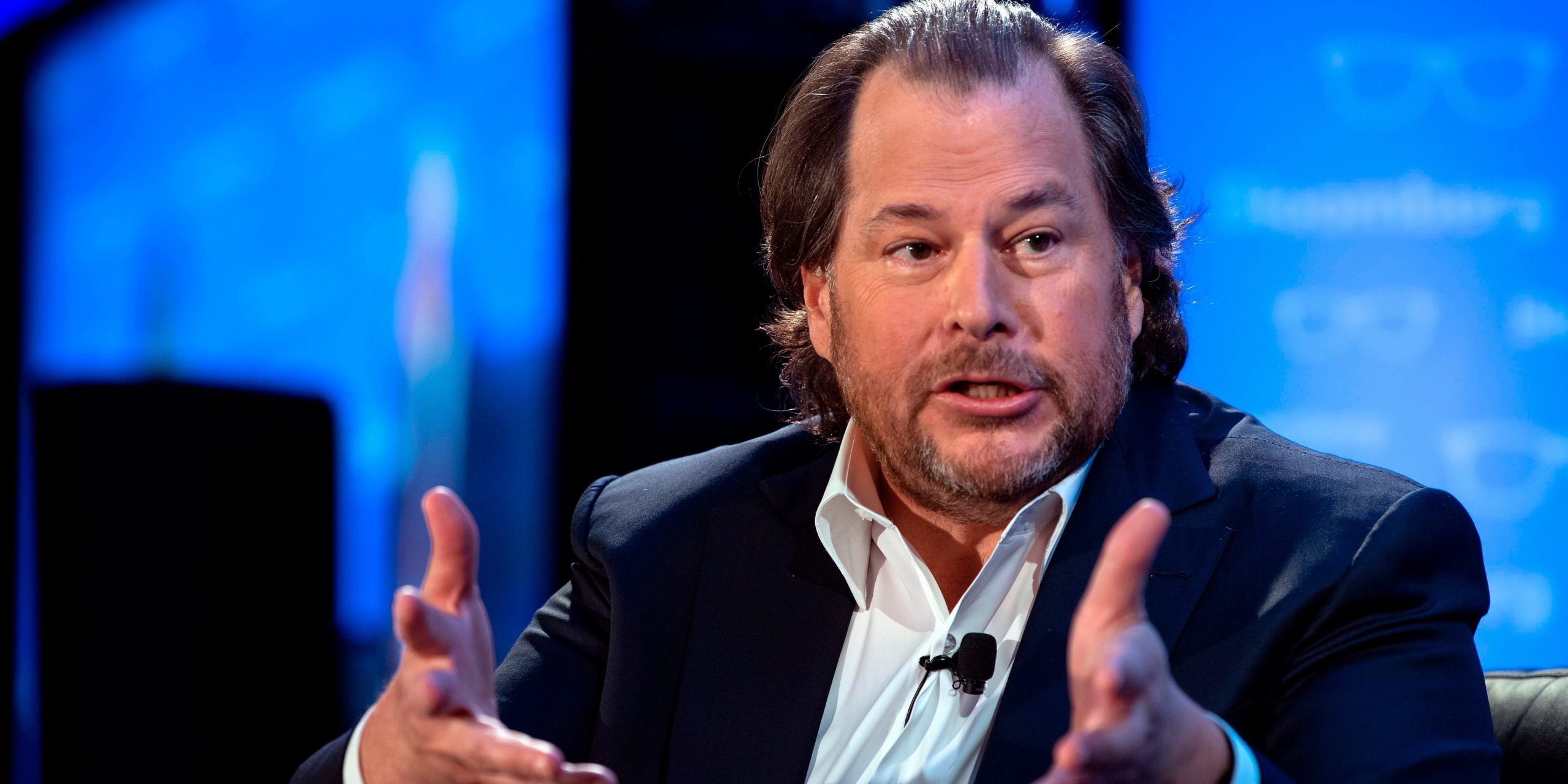 Salesforce founder Marc Benioff, wearing a sports coat jacket and in front of a blue background on a stage, extends his hands forward during a talk.