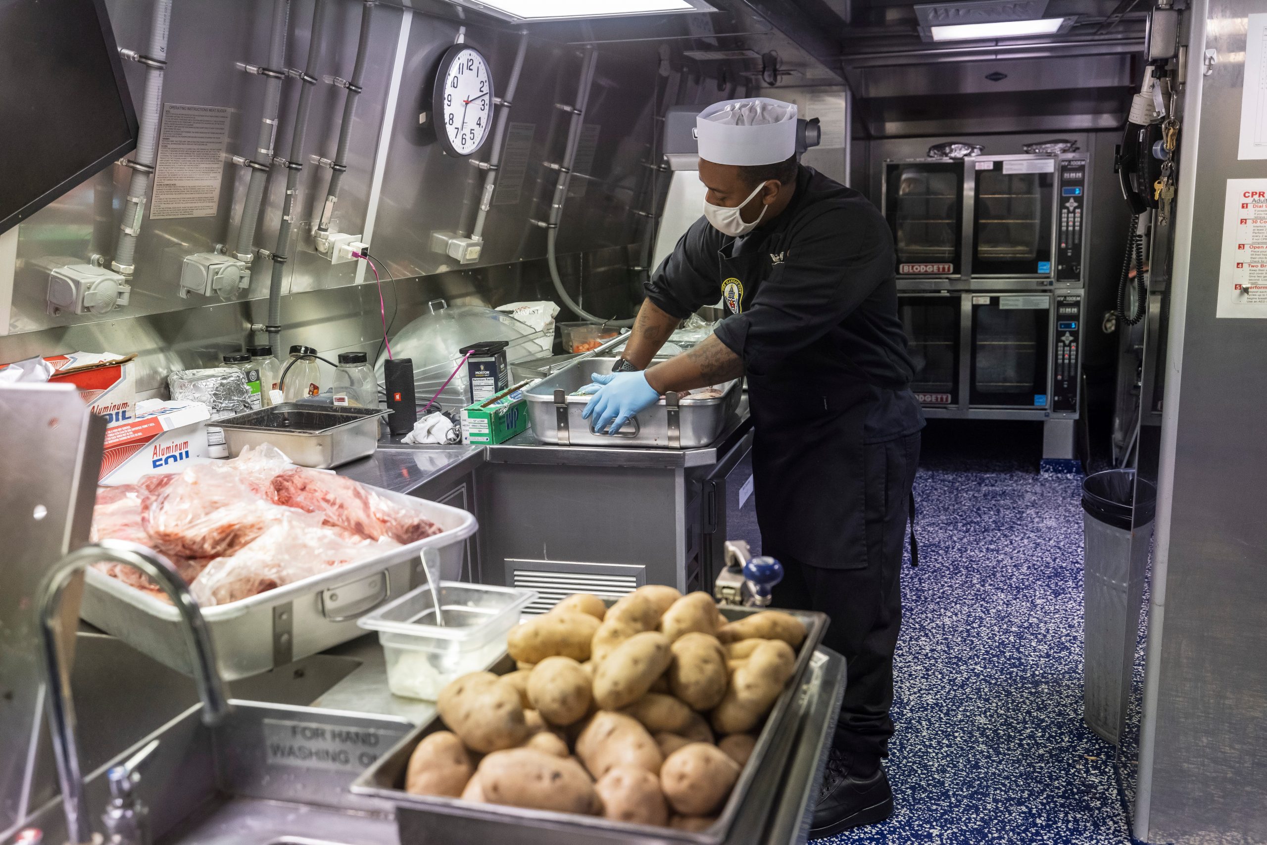 A man wearing a mask is seen preparing food with containers in the foreground.