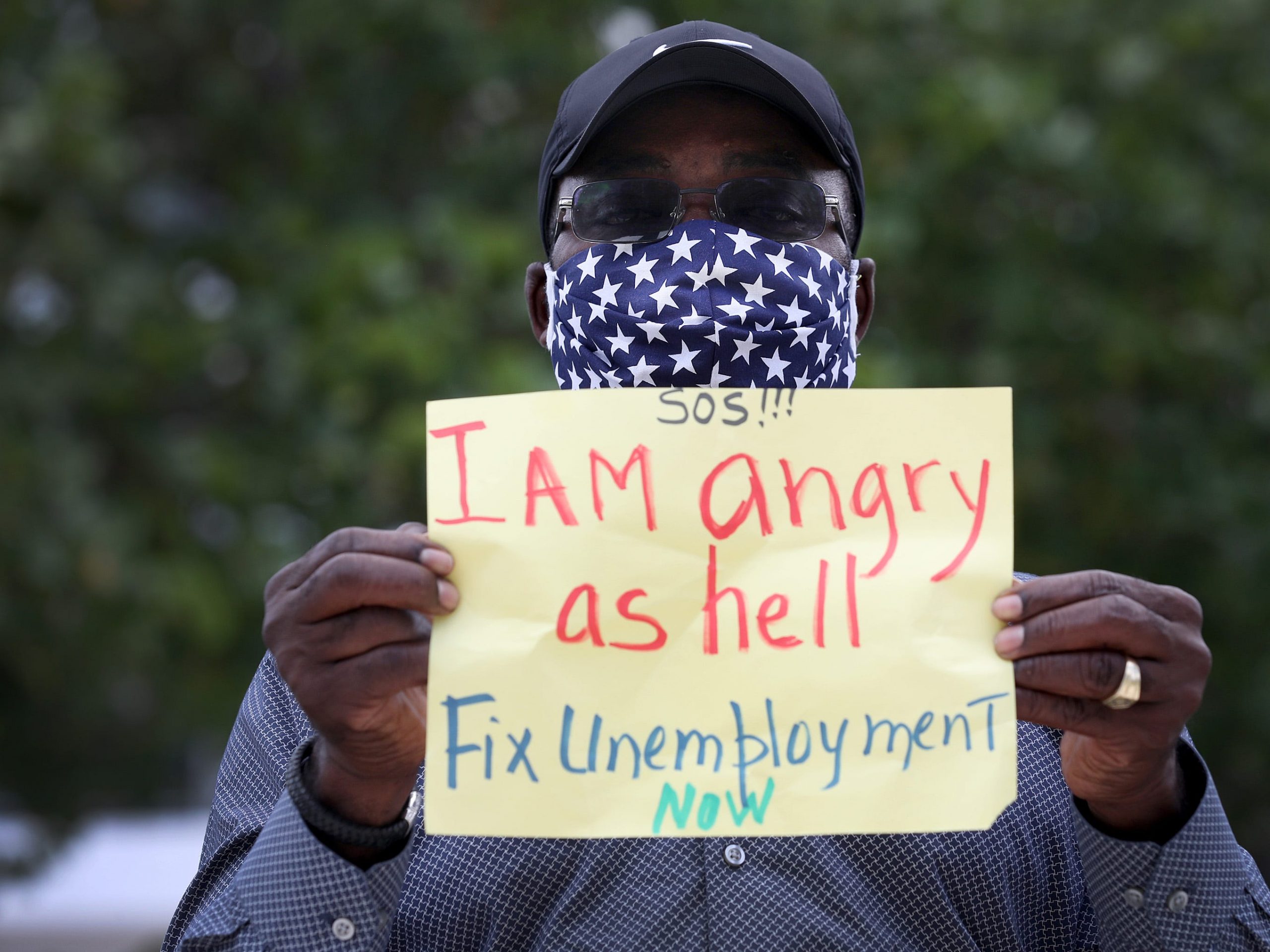 an unemployed worker holds a sign that says I Am angry as hell Fix Unemployment Now,'