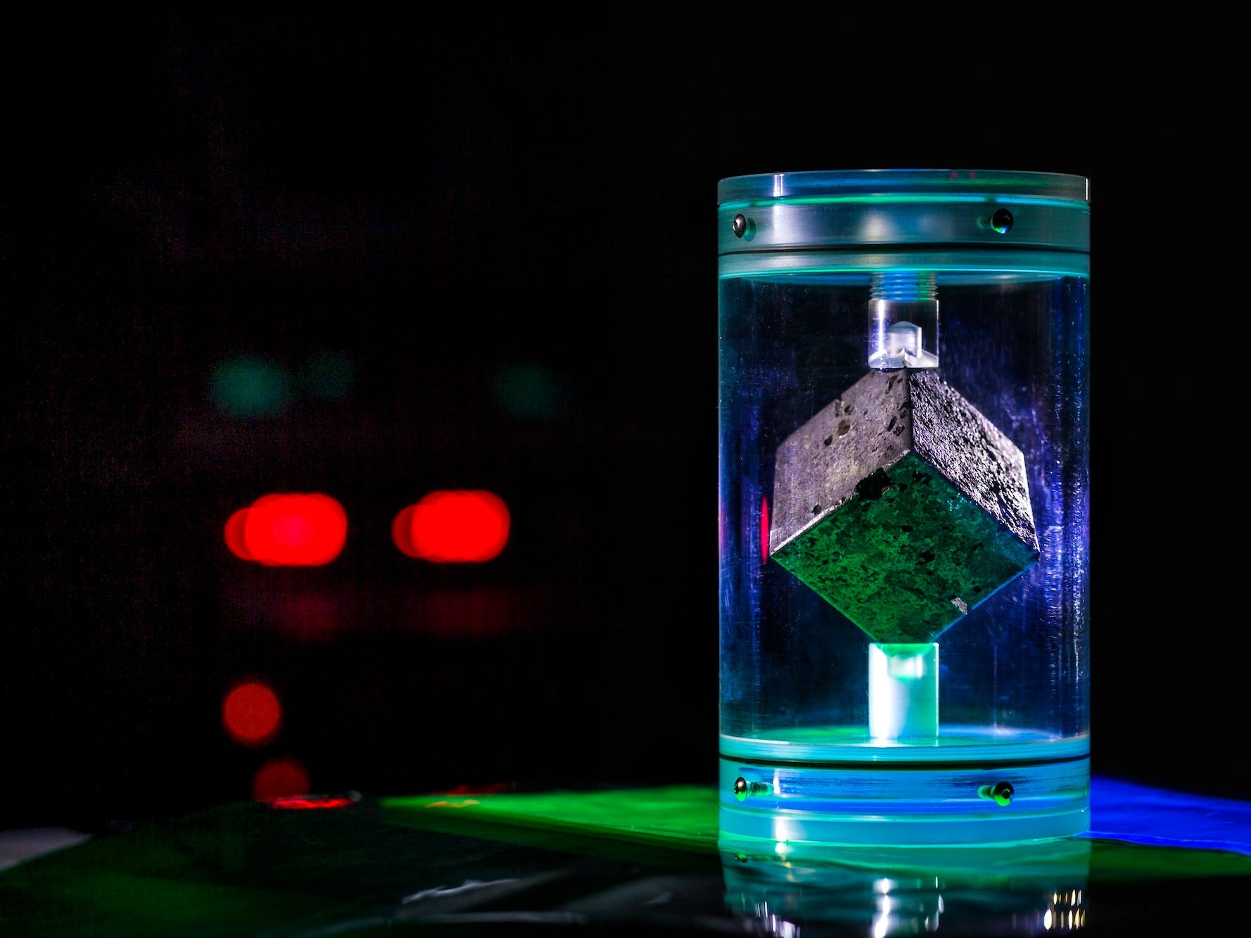 a grey cube is balanced on its vertices in a transparent display casing, in green and blue light