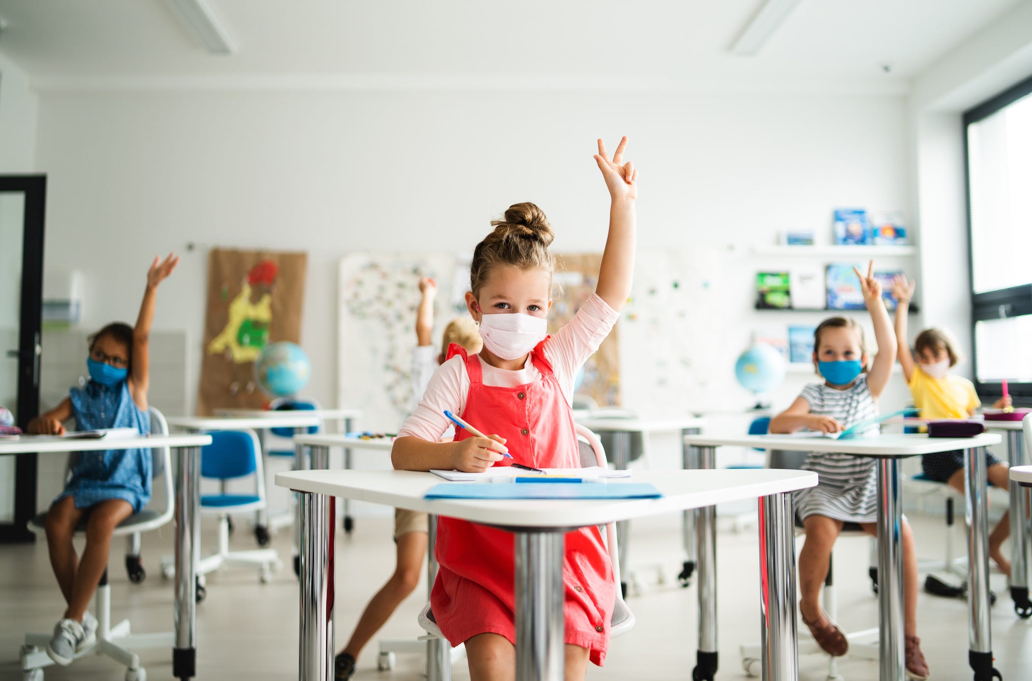 A student in a socially distanced classroom raises her hand, with a mask on.