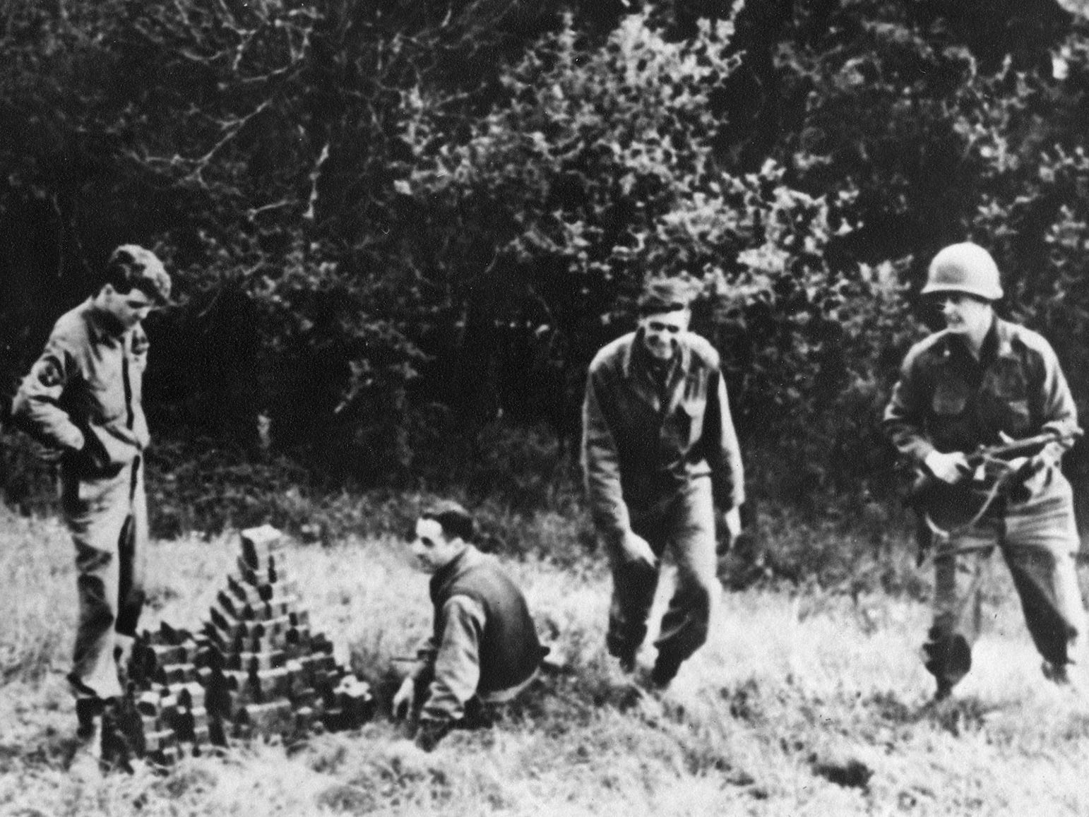 A black and white picture shows men in uniform around a hole in the ground, one is sitting, in the center of the image is a pile of cubes, in the background are trees.