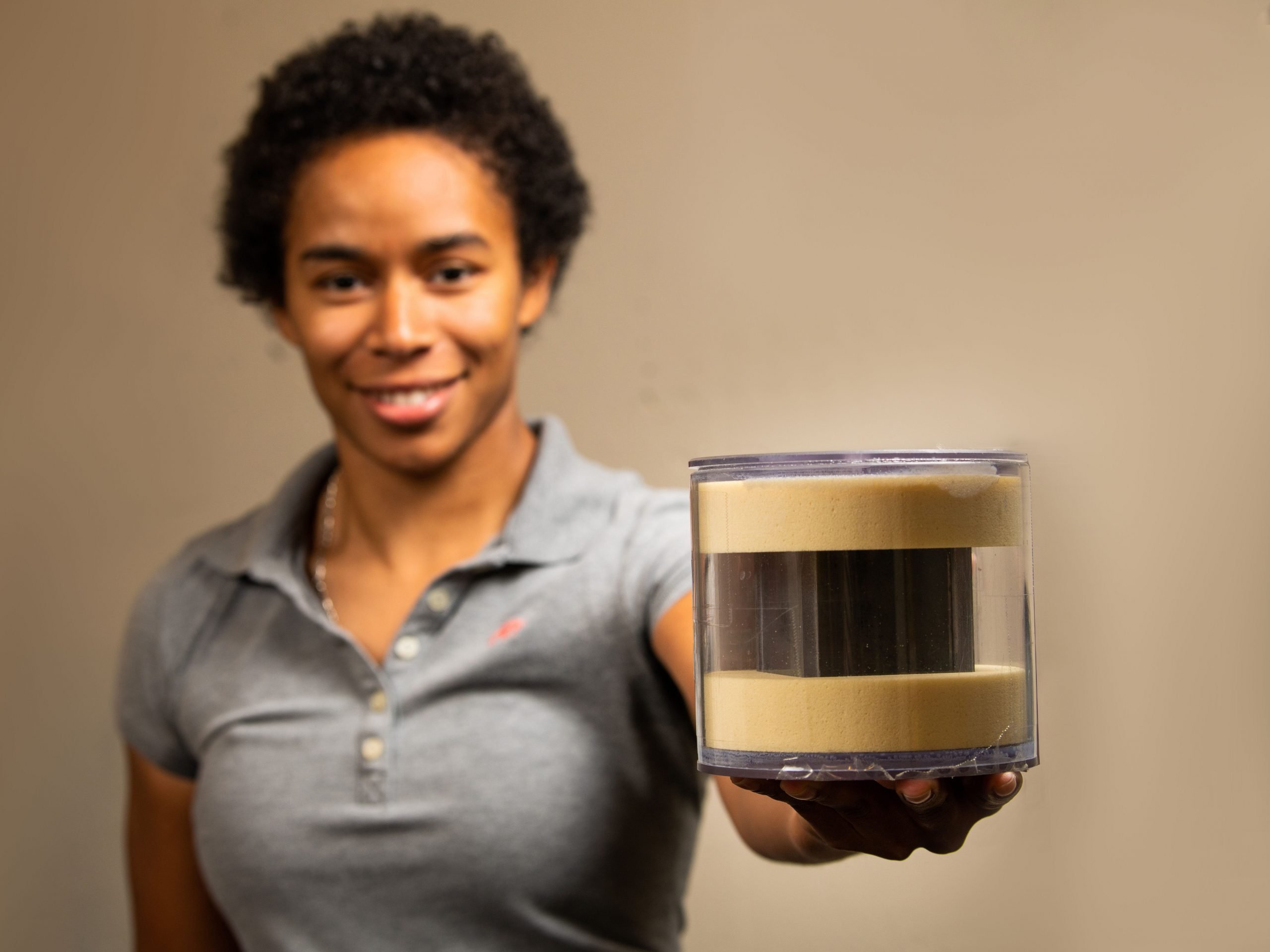 A woman wearing a grey shirt holds out a circular plastic container which contains a grey cube encased in padding