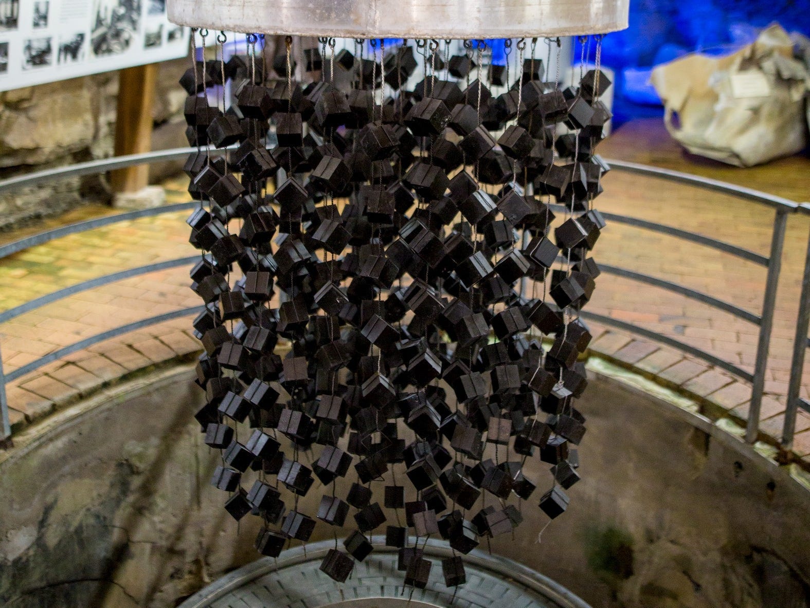 grey cubes are hanging from a silver metal plate above a cylindrical hole in the ground.