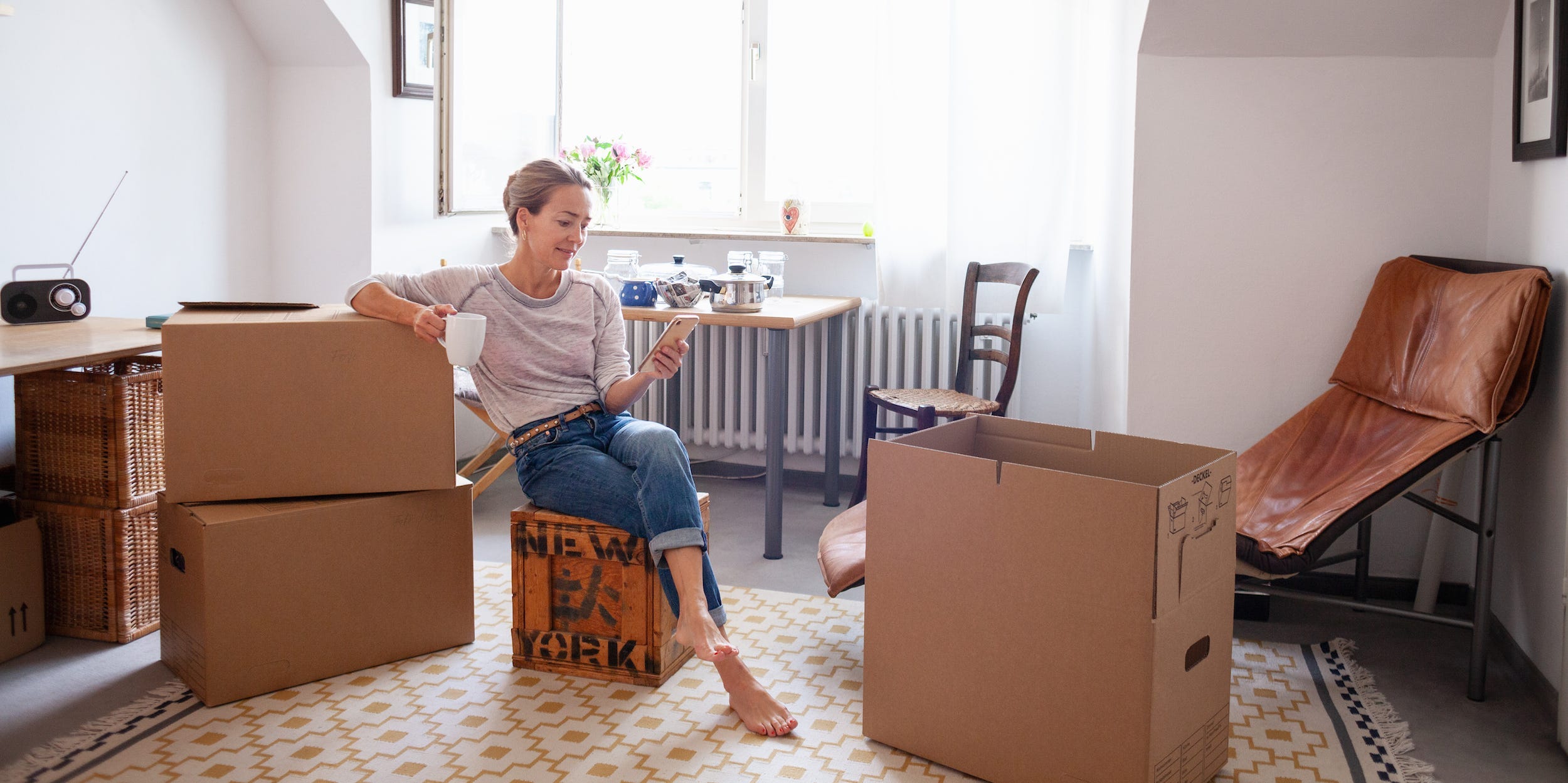 woman packing up apartment with boxes around her, holding smartphone