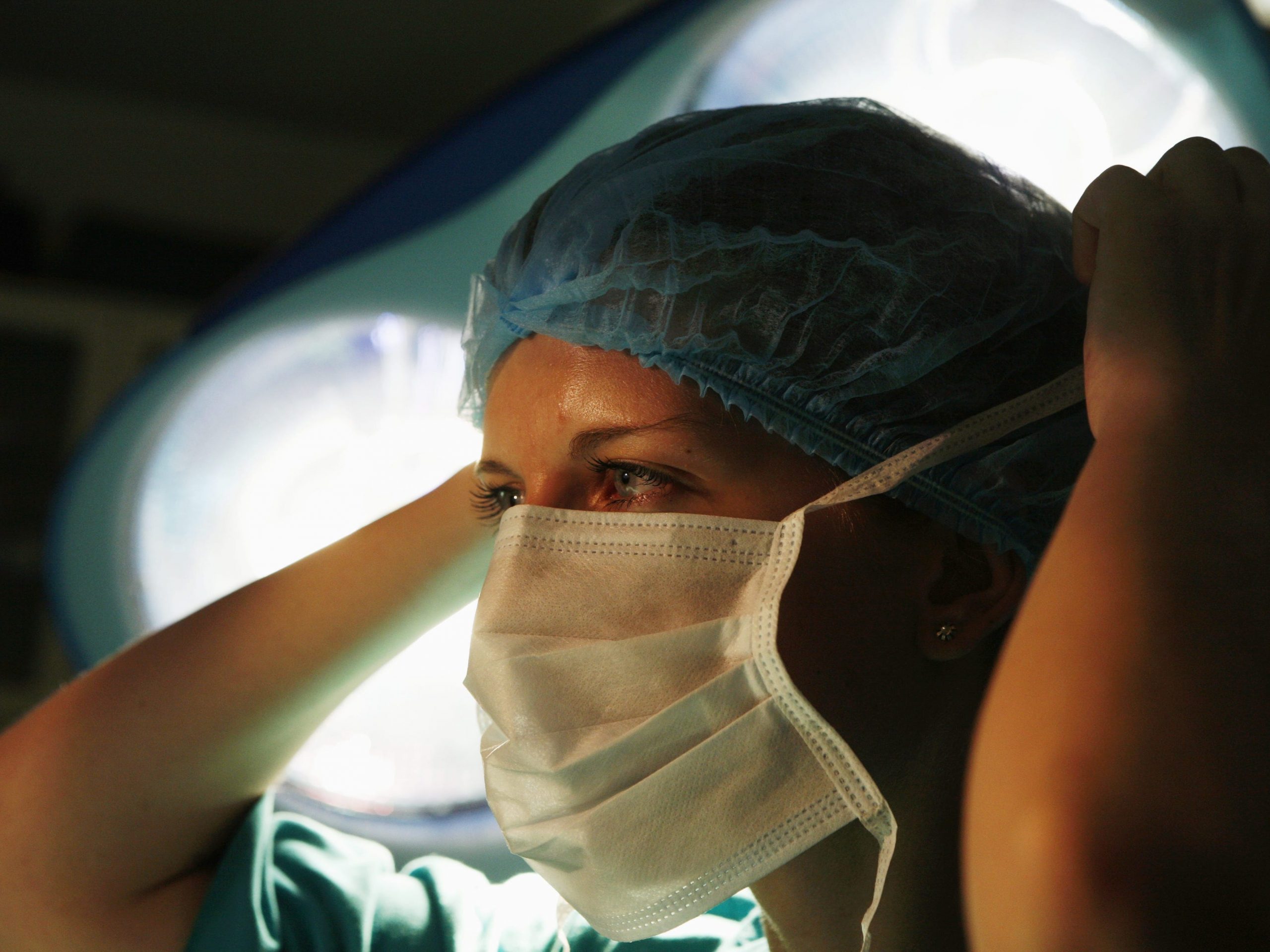 Female surgeon ties her surgical mask around her face underneath operation lights.