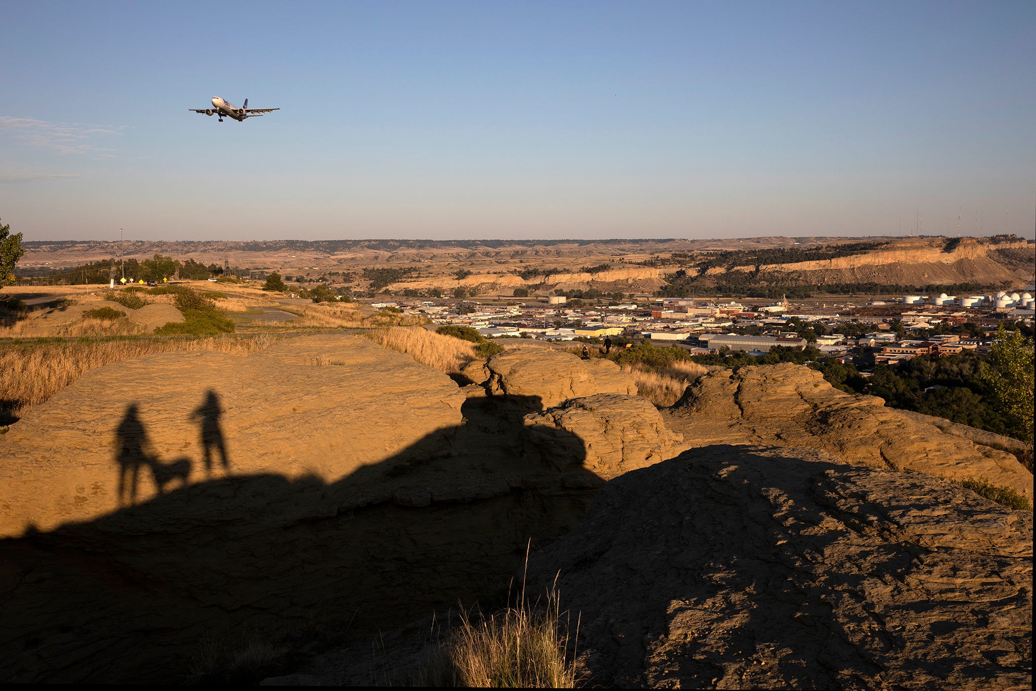 A plane flies over orange rocks that have the shadows of two people and a dog cast against them.