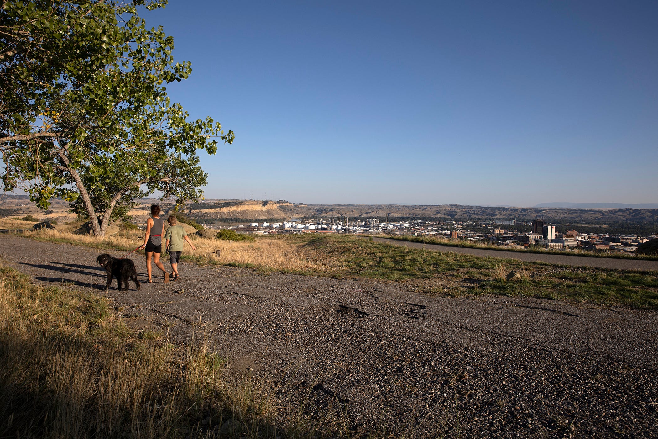 Two people walk their black dog along a hiking trail which overlooks Billings, Montana under a dark blue sky.