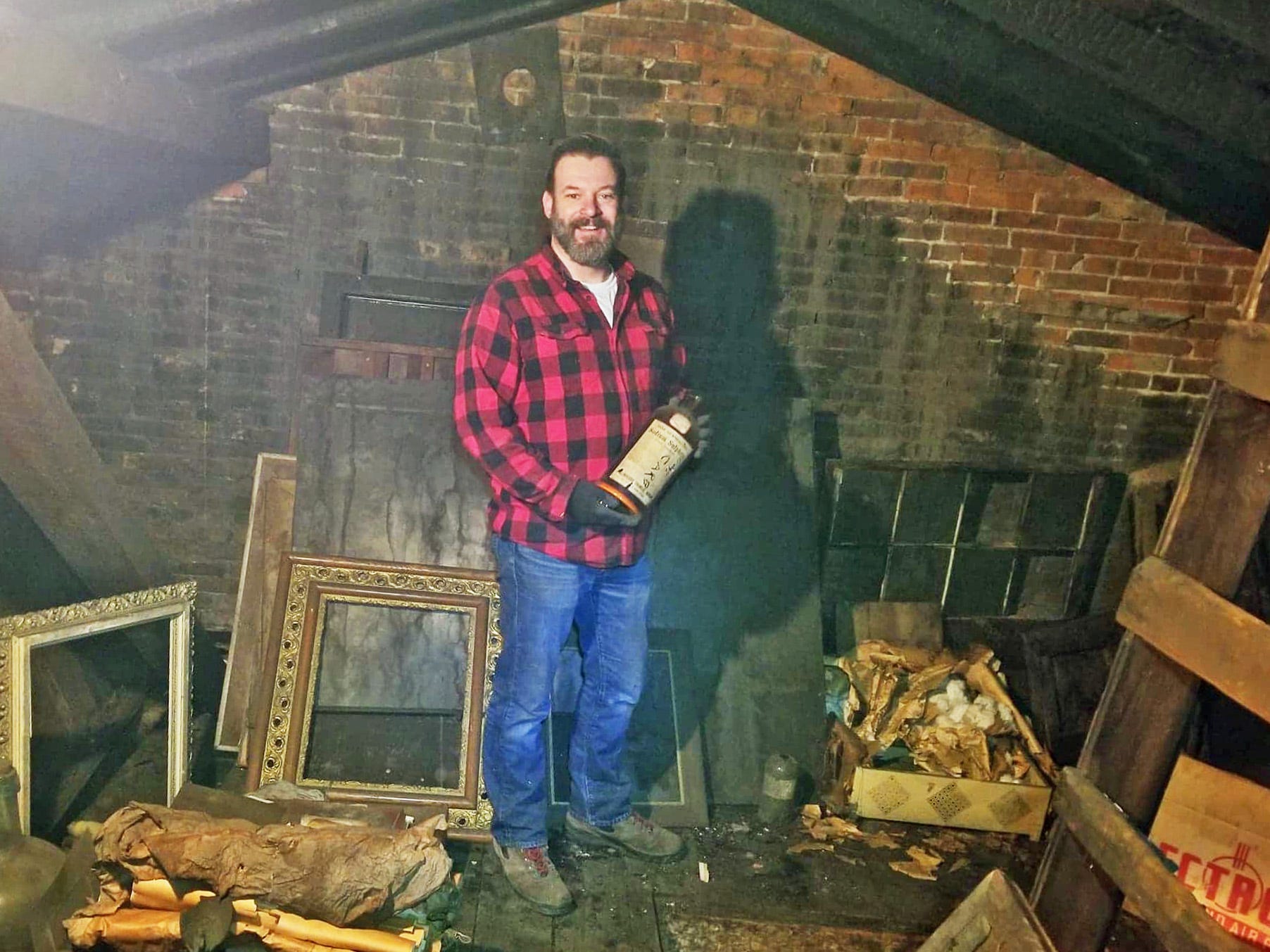 A man in a red flannel and blue jeans stands smiling in a dimly lit attic filled with antiques