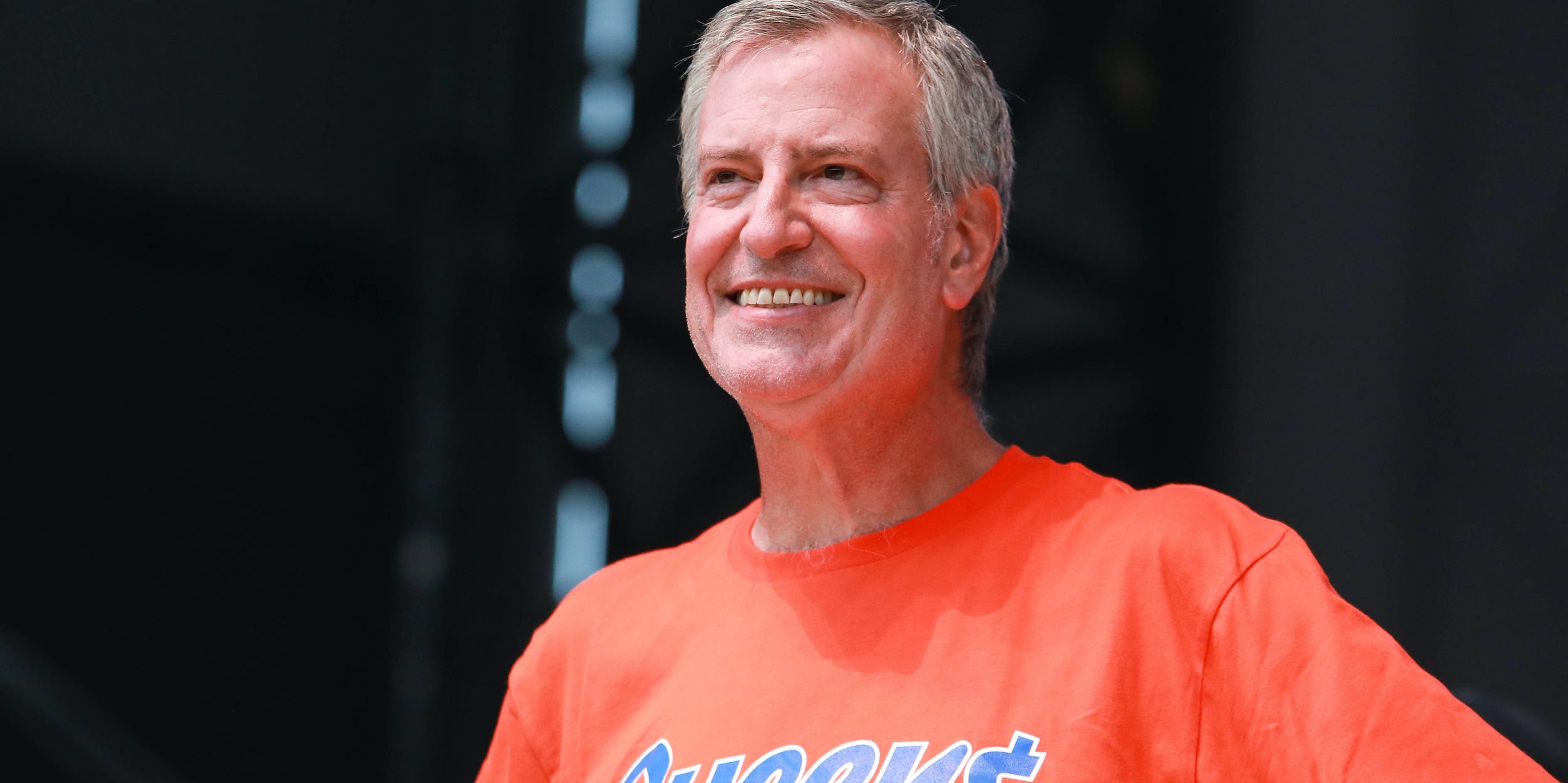 New York City Mayor Bill de Blasio stands with his hands on his hips in an orange "Queens: Get the Money" t-shirt.