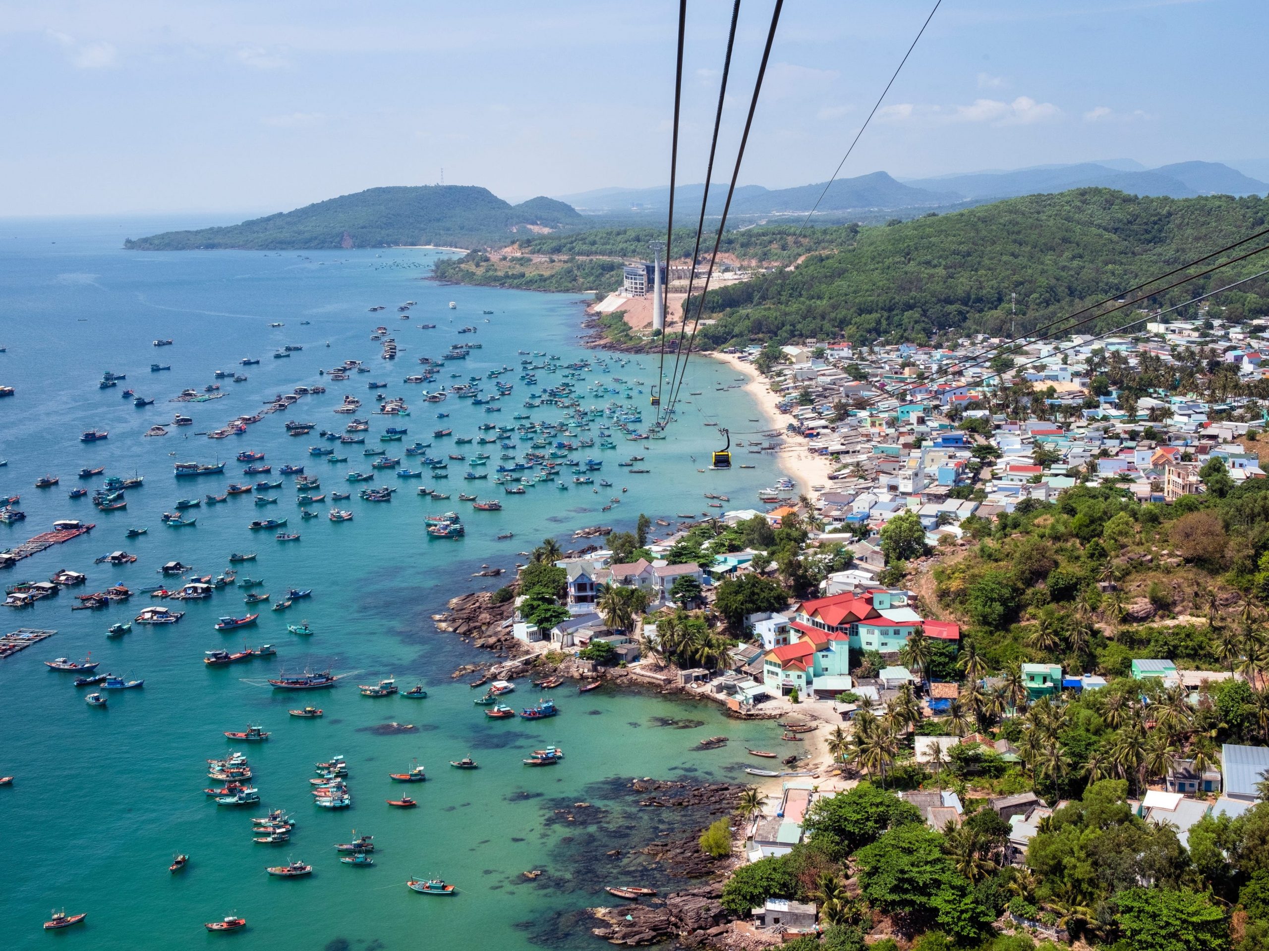 An aerial view of an island shows fishing boats in a bay and a cable car stretching across the beach