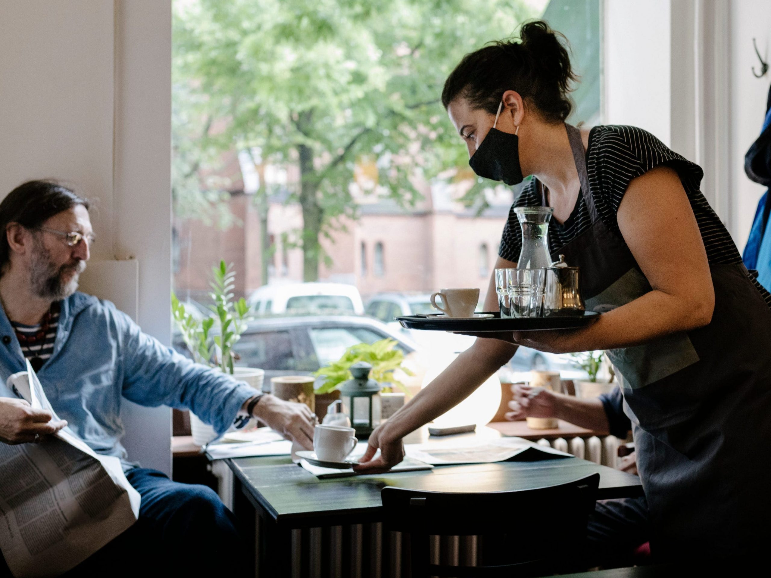 A waitress wearing a face mask and serving a customer some coffee at his table in a restaurant.