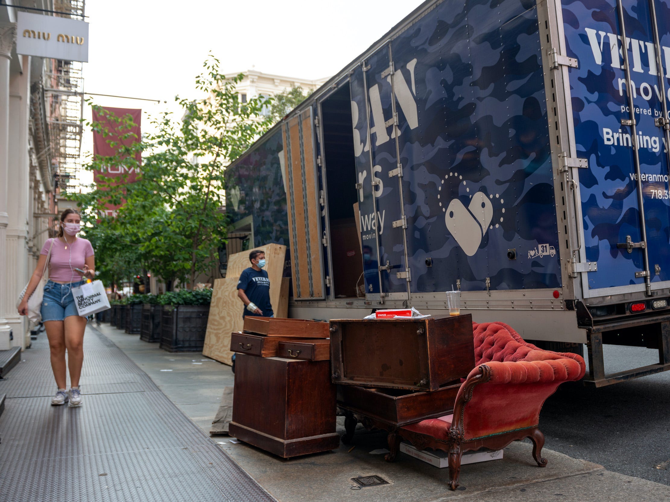 Woman in mask walks past moving truck and furniture on street