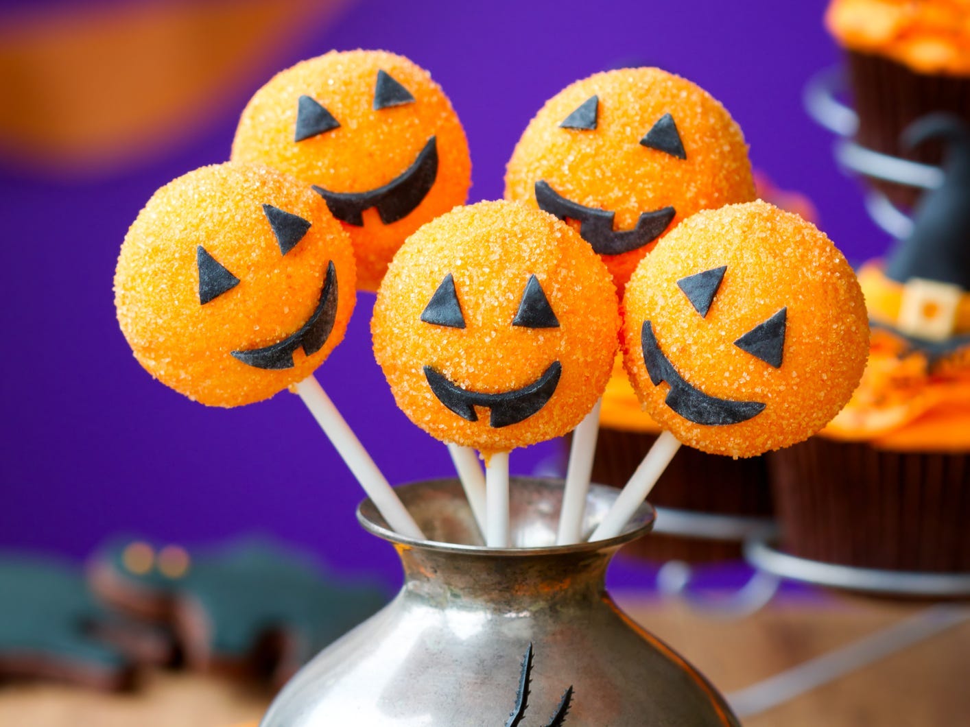 Cake pops decorated like jack-o-lanterns displayed in a silver urn with a plastic spider on it