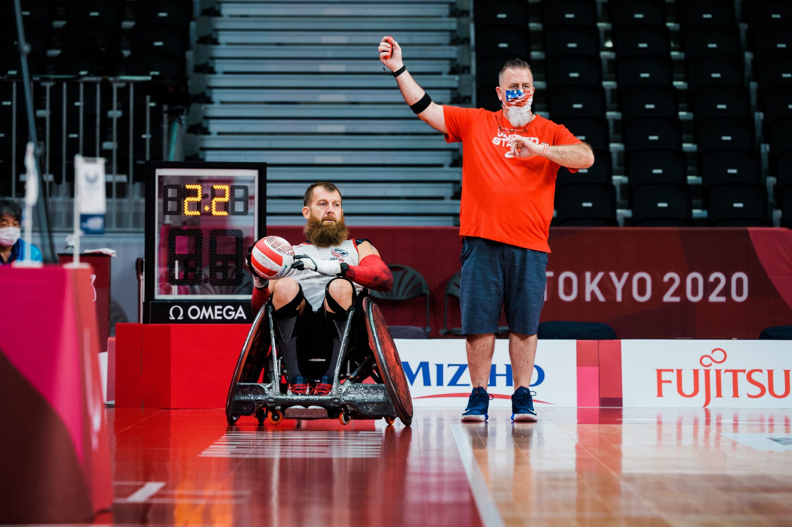 Wheelchair rugby player sits holding a ball next to standing man in mask holding his right arm up