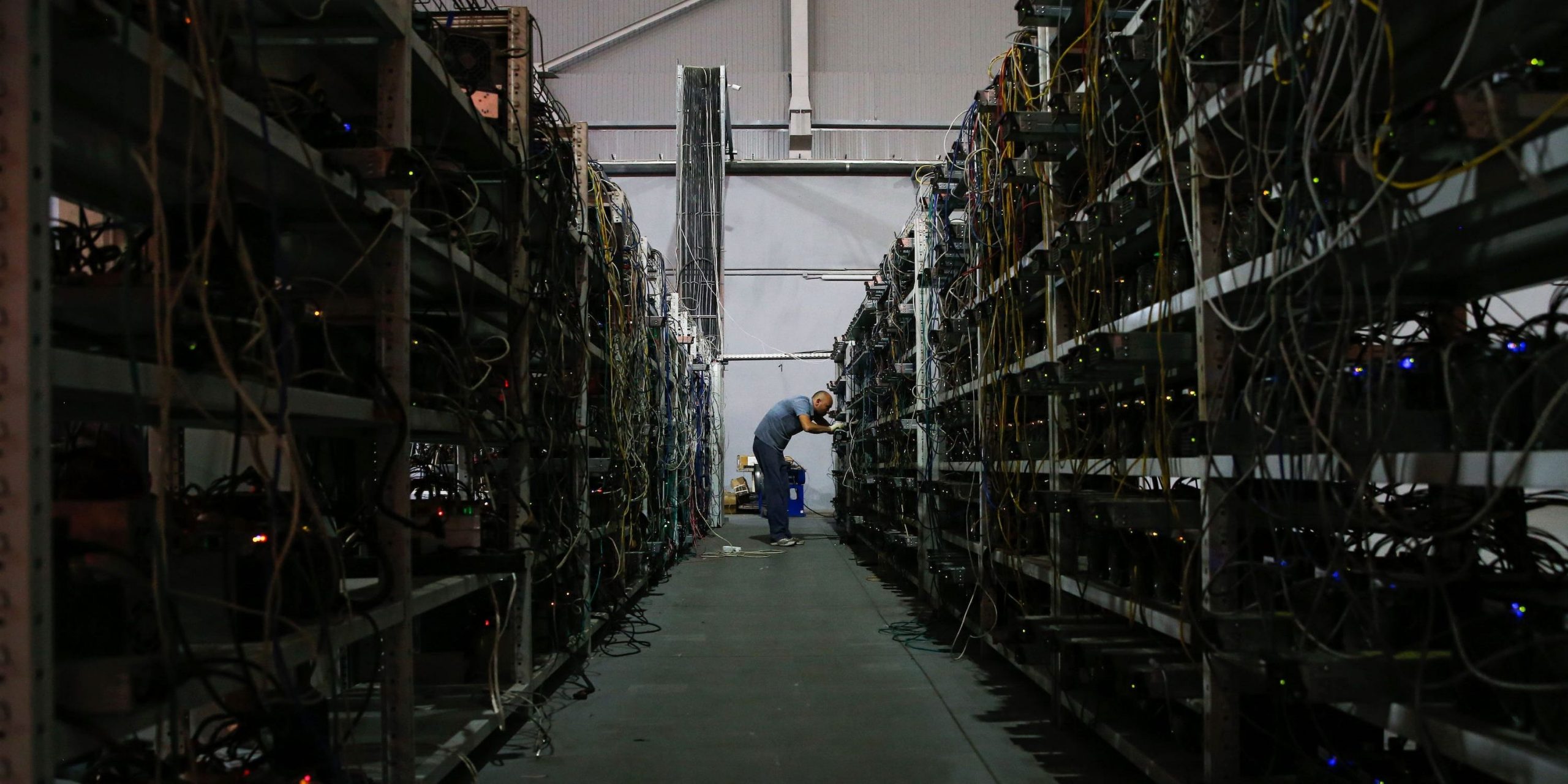 A man stands at the end of a long corridor. The walls are packed with computers and wires, which are being used to mine bitcoin.