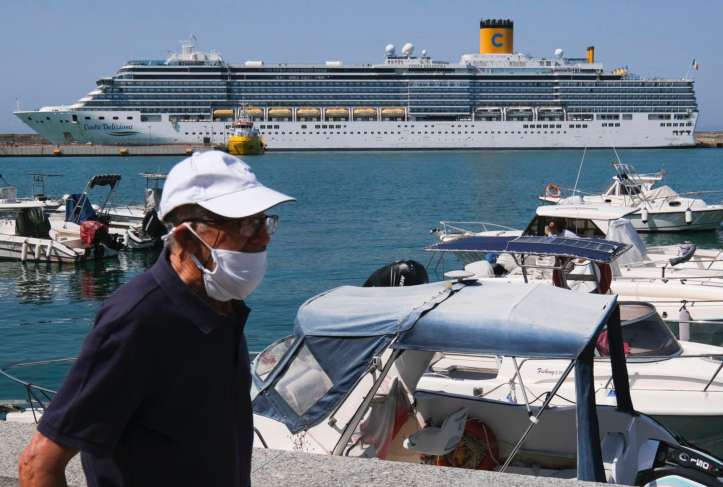 A white cruise line sits in the ocean in Italy in front of smaller boats and a man wearing a mask and a white baseball cap.