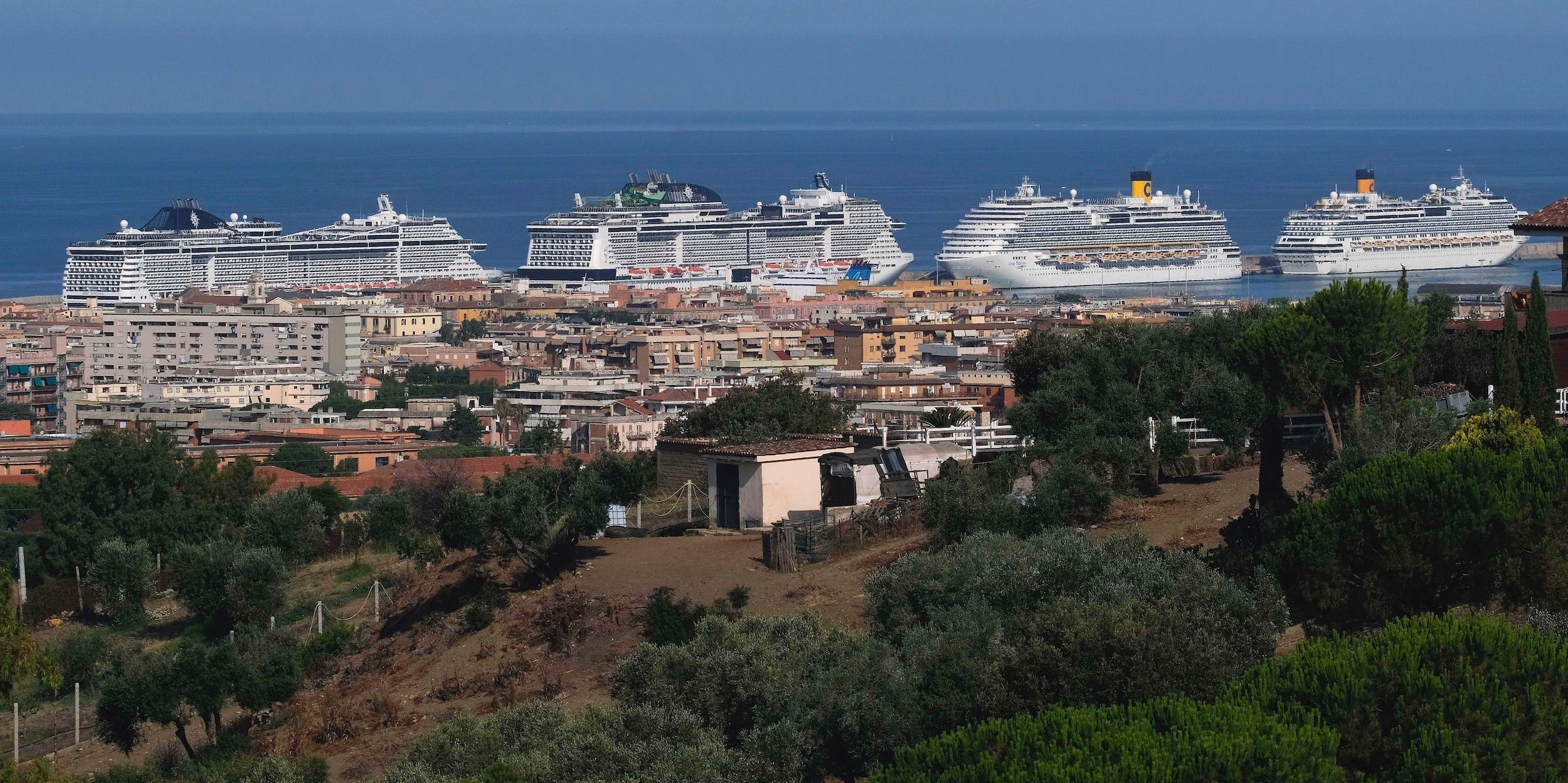 White cruise lines float in the ocean in front of buildings a hill that has bushes on it.