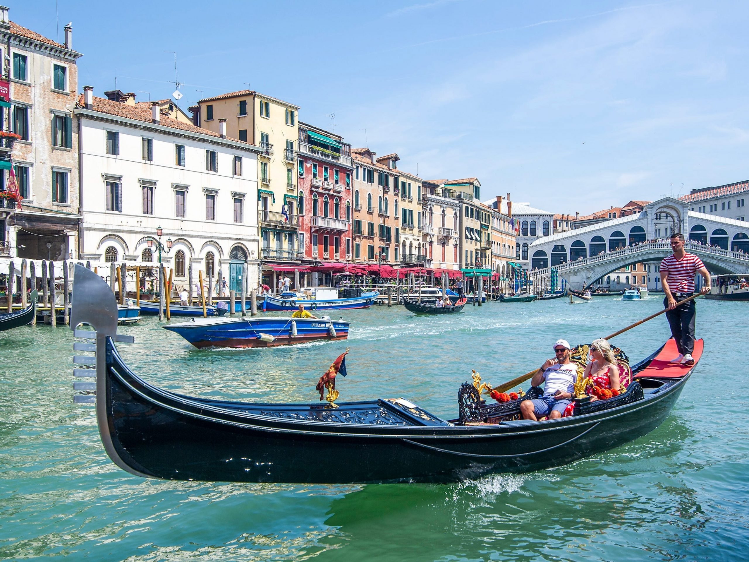 Tourists sit in a gondola boat in Venice, Italy, as a gondolier steers the boat surrounded by colorful buildings and bright-blue water.