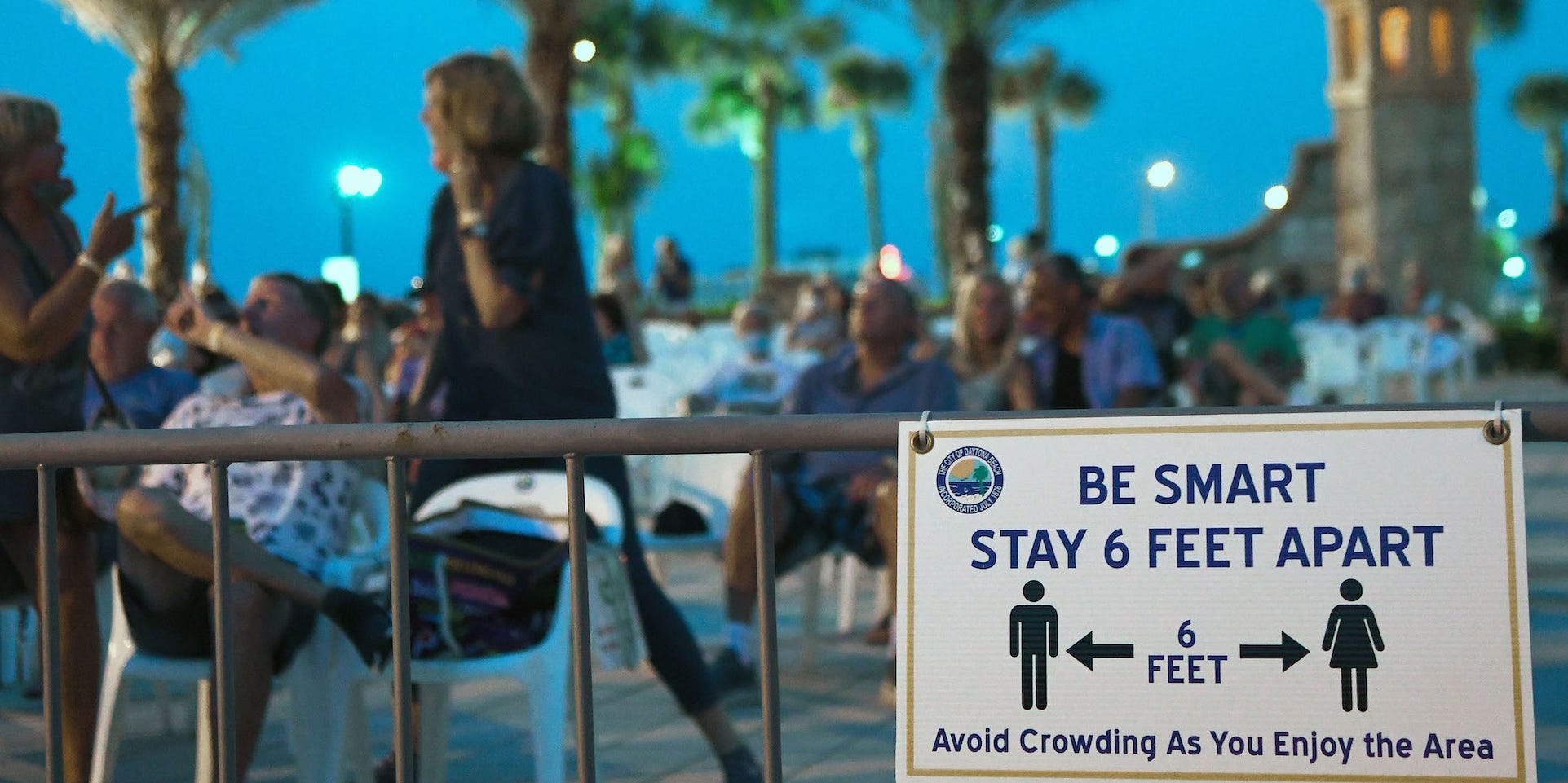 People at night outside behind a gate that reads "be smart, stay 6 feet apart"