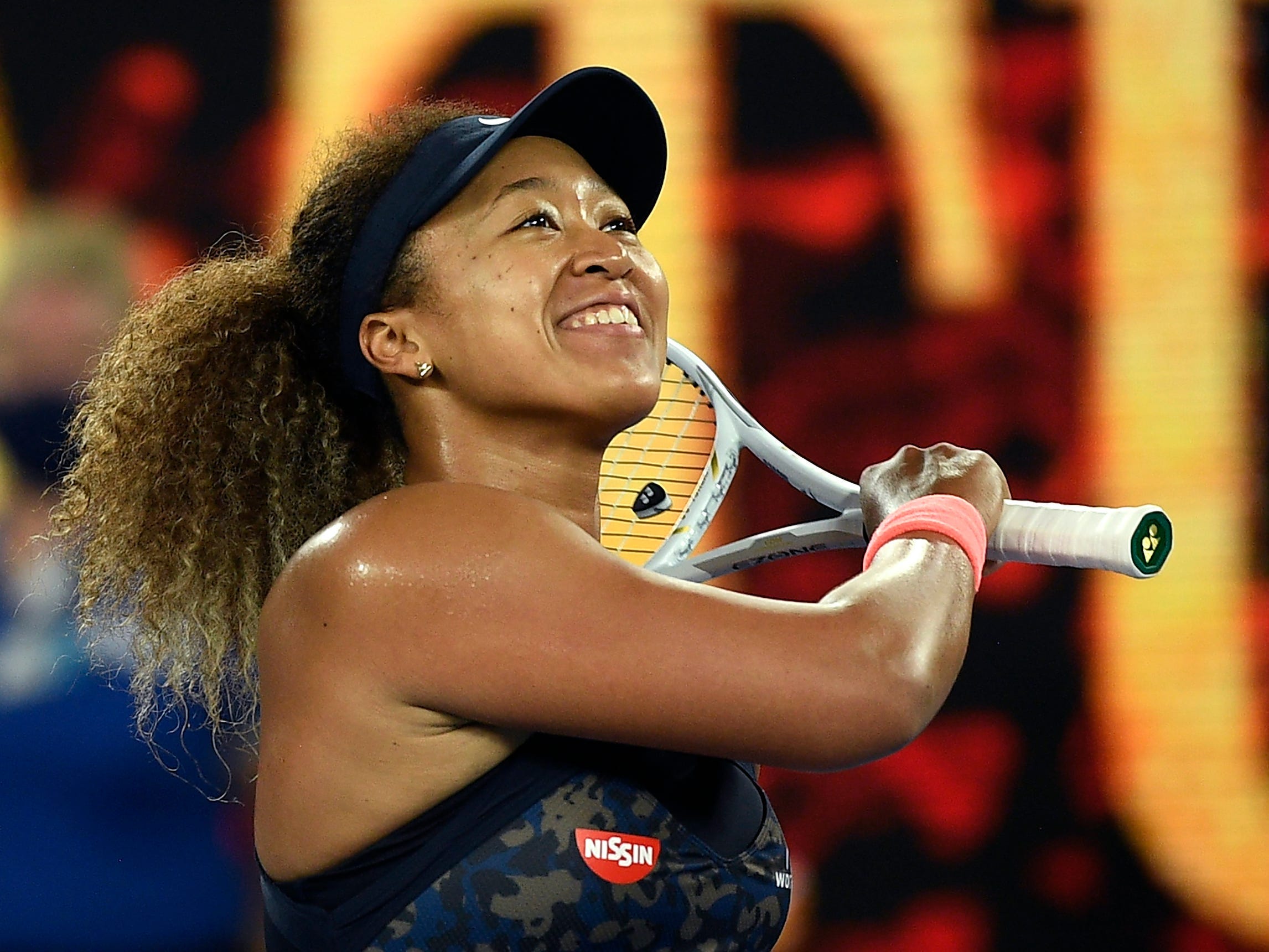 Naomi Osaka smiles while wearing a visor and holding a tennis racket over her shoulder