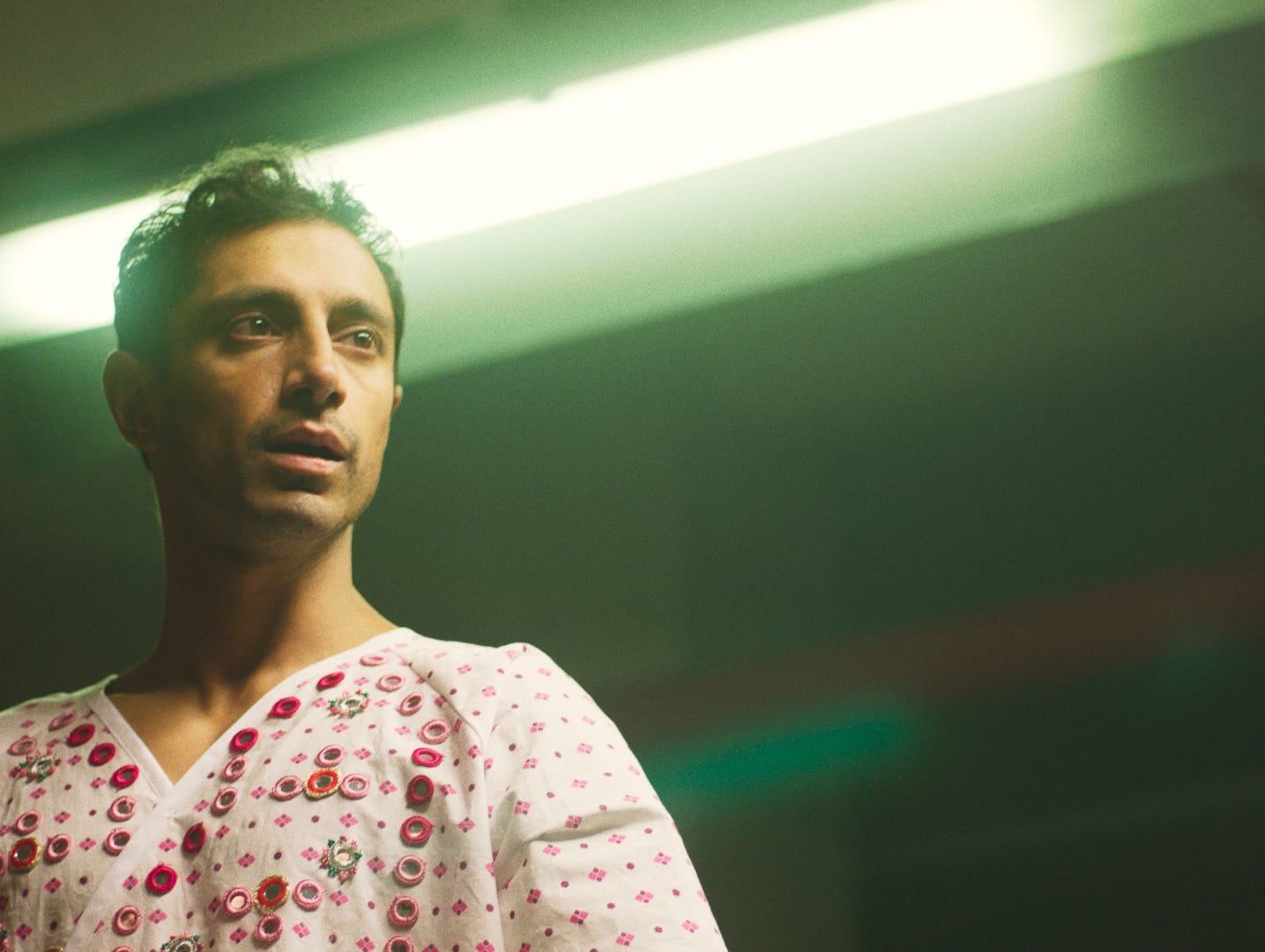 Actor Riz Ahmed in the movie "Mogul Mowgli," standing in front of a flourescent light while wearing a hospital gown.