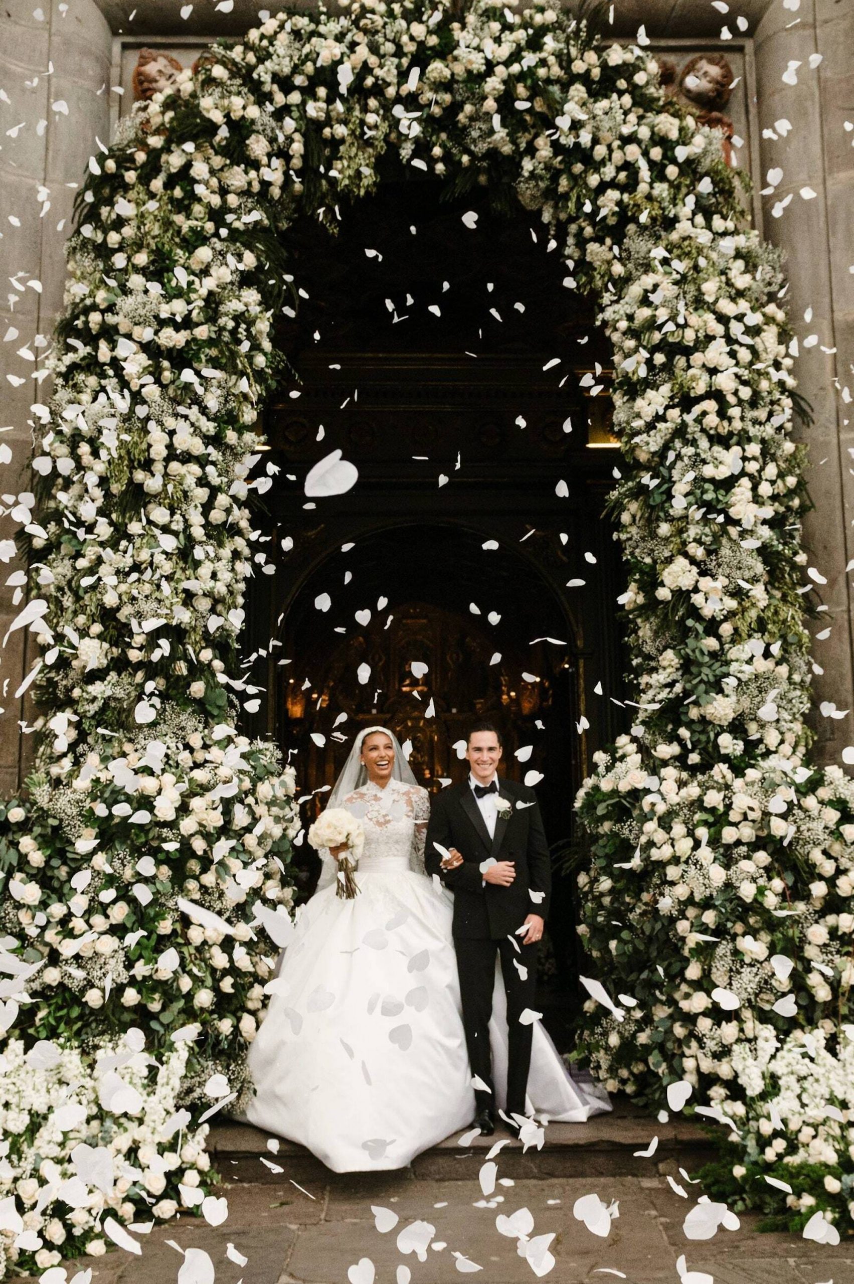 A bride and groom stand under a floral archway as flower petals fall around them.