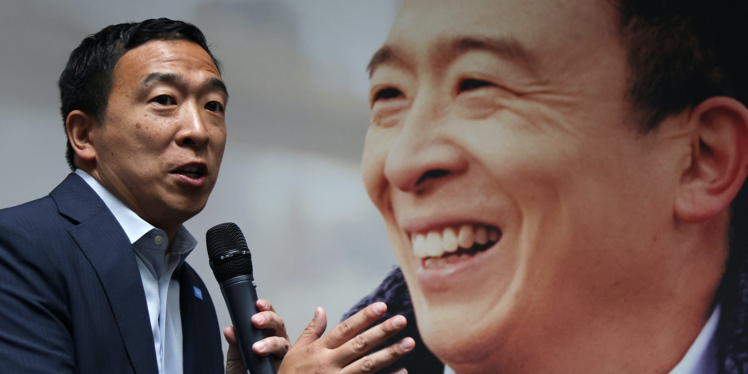 Andrew Yang speaks with a microphone in front of a blown up portrait of his face.