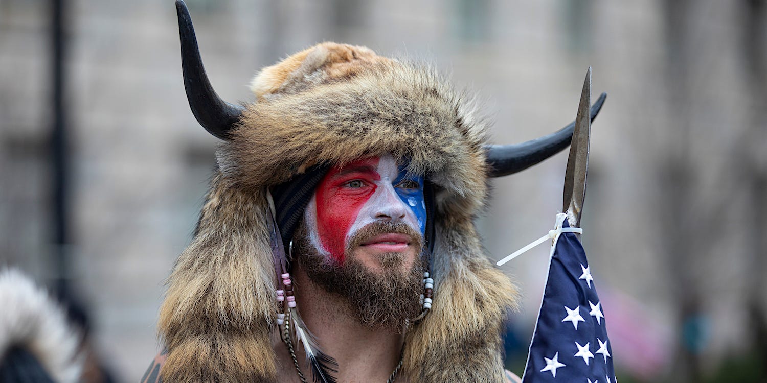 A shirtless man with red, white, and blue face paint, a fur head covering with two horns holding an American flag with a spear tip