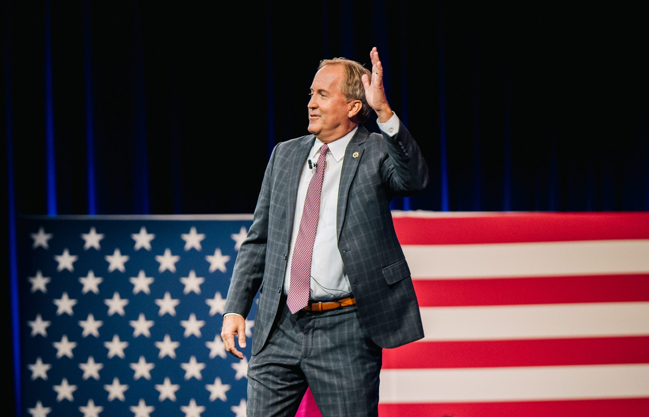 Texas Attorney General Ken Paxton waves in a suit in front of an oversized American flag