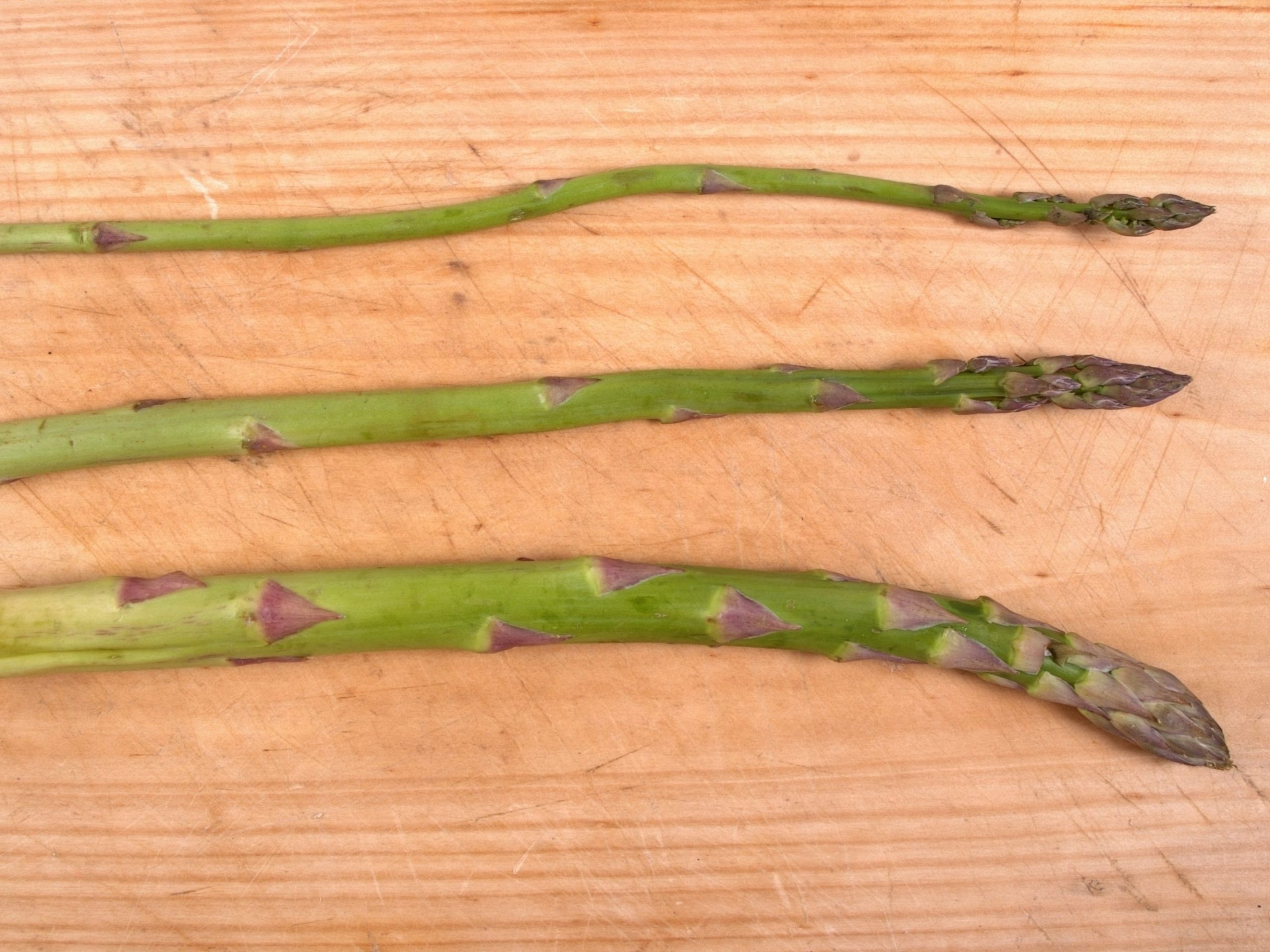 Three asparagus stalks on a cutting board. The top one is very skinny, the middle one is standard size, and the bottom one is thick.