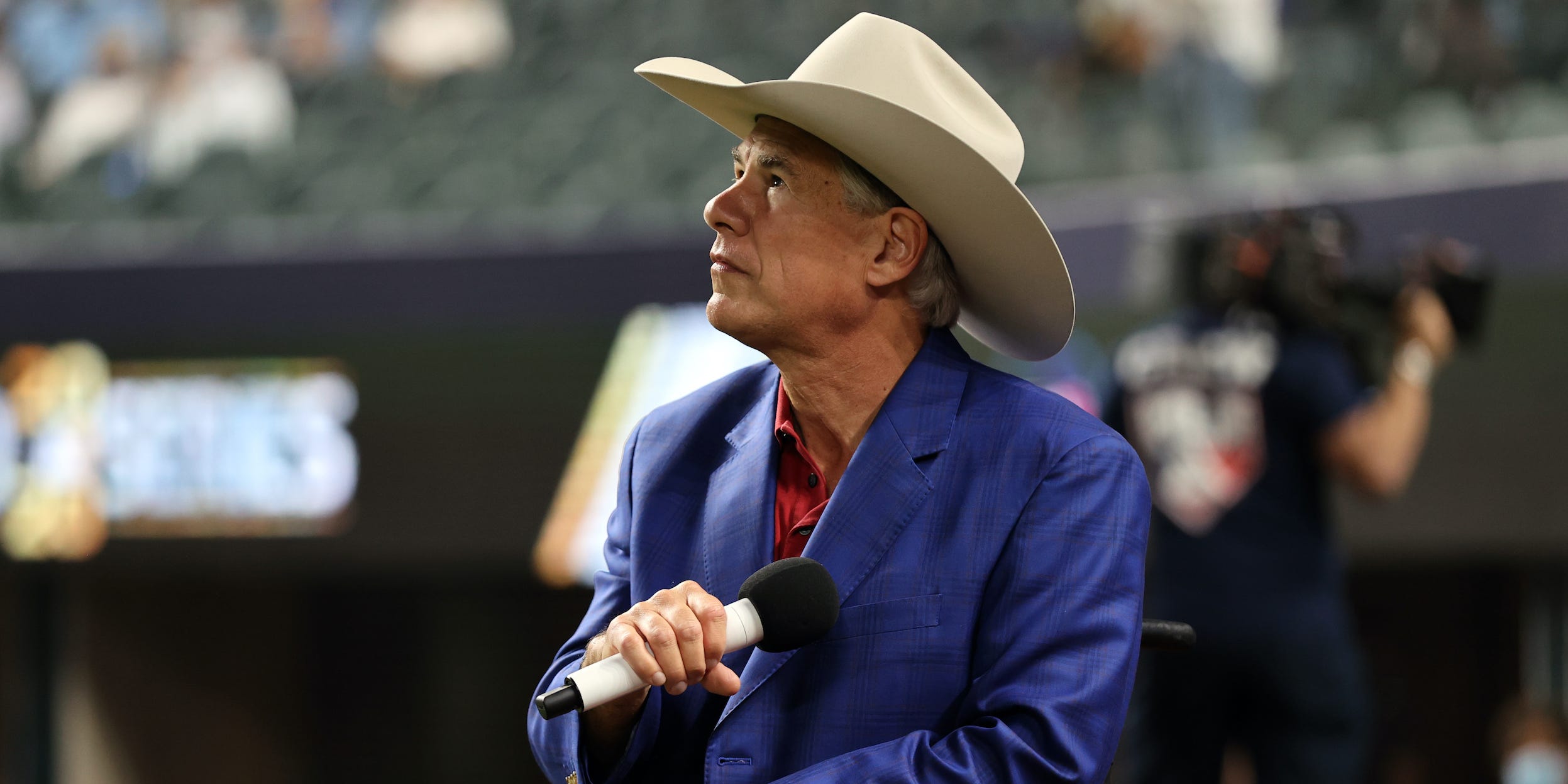 Wearing a tan cowboy hat, Texas Governor Greg Abbott lowers a microphone toward his chest while listening to the national anthem at a World Series baseball game held in Arlington, Texas.