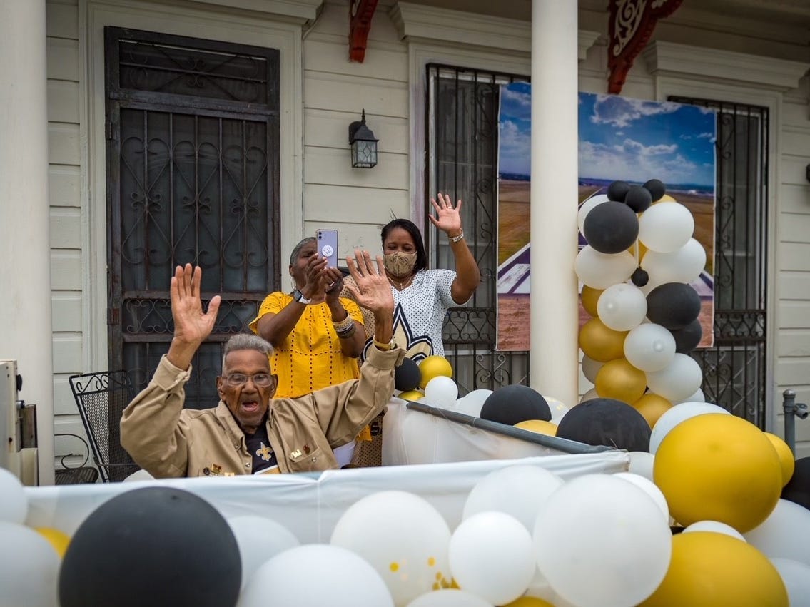 Lawrence Brooks raises his hands above his head and smiles as he celebrates his 112th birthday on his front porch, surrounded by white, black, and gold balloons.