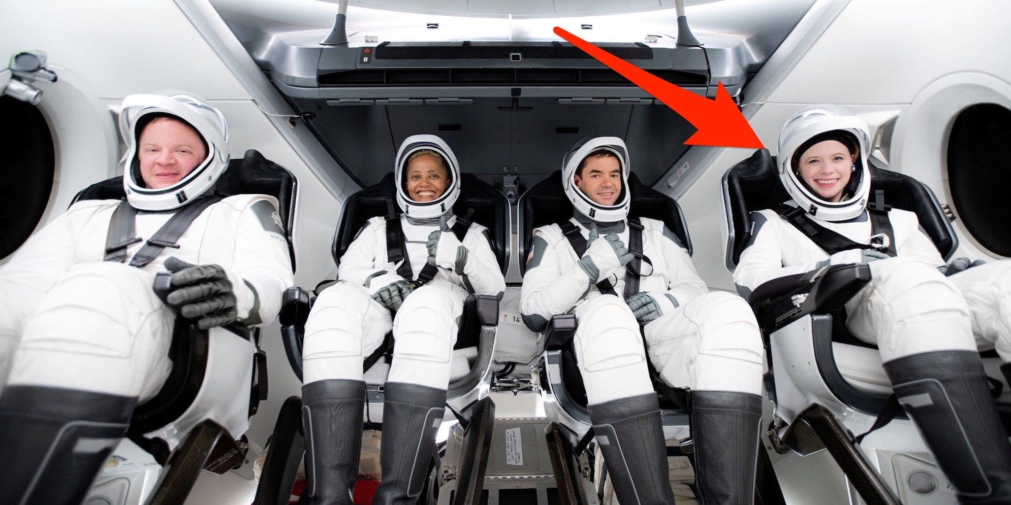 A picture of the SpaceX Inspiration 4 crew in a model Crew Dragon spaceship, smiling and they are sitting in their chairs and wearing they full black and white spacesuits. A large red arrow points to Hayley Arceneaux.