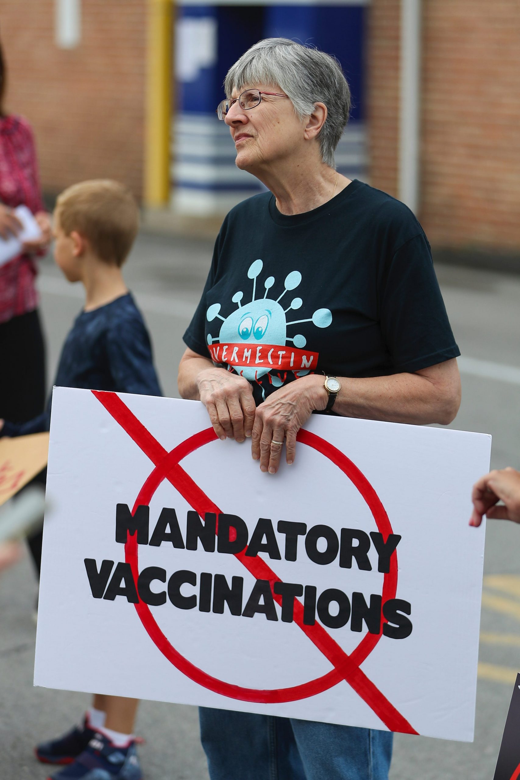 woman at vaccine protest standing holding sign ('no mandatory vaccinations') is wearing FLCCC shirt promoting the use of ivermectin, a deworming drug that has not been proven to help patients with COVID-19.